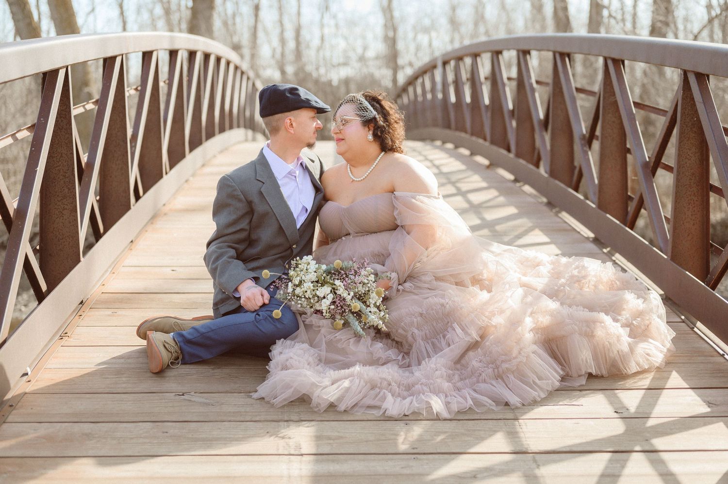 Bride in blush tulle gown and groom in gray suit sitting together on a wooden bridge.