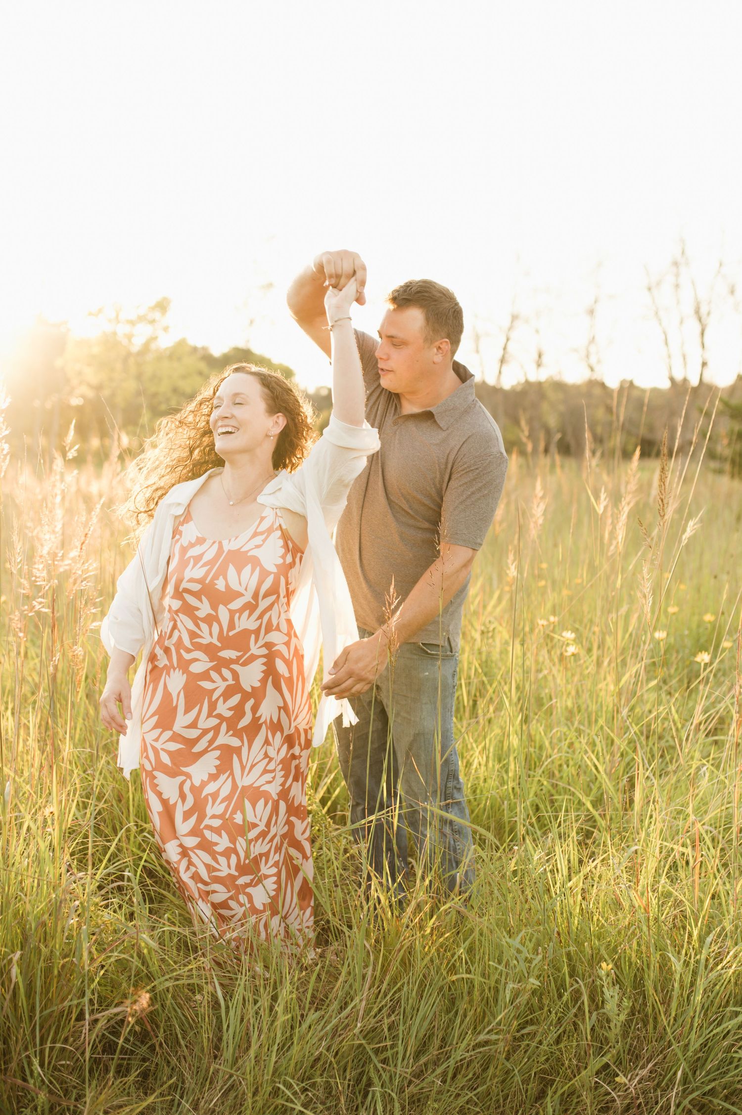 A couple dances joyfully in a sunlit meadow, the man spinning the woman in a floral orange dress.