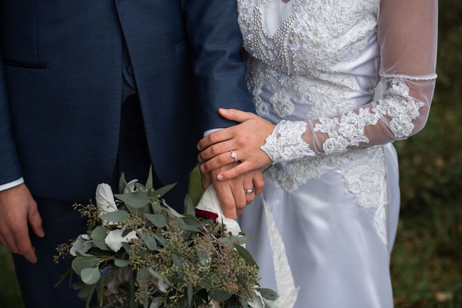 Close-up of bride and groom holding hands showing wedding rings with bridal bouquet of eucalyptus.