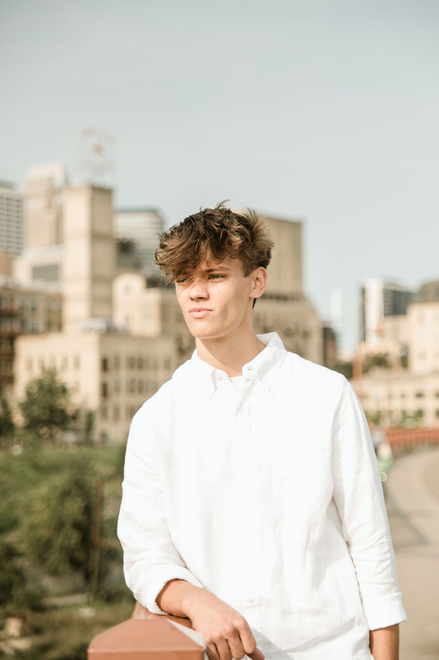 Teen boy in white linen shirt standing on rooftop with city skyline background.