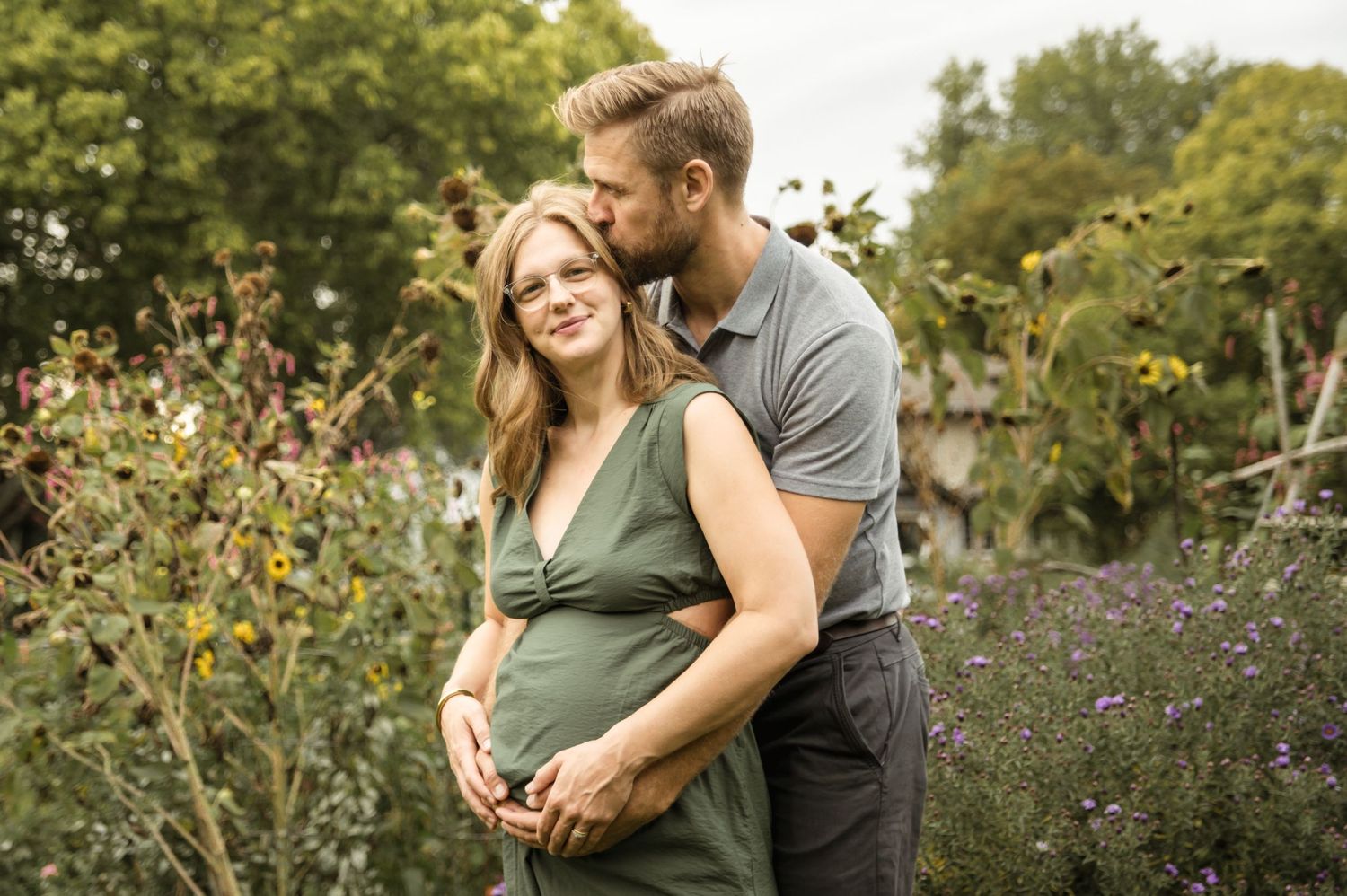 Couple sharing tender moment outdoors, man kissing pregnant woman's head while cradling her baby bump in garden.