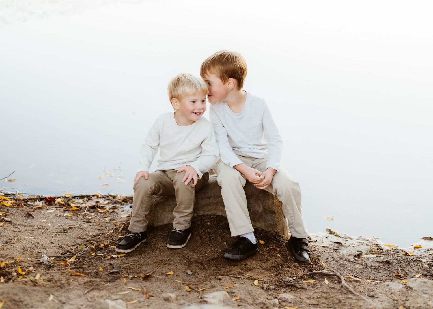 Two young brothers sitting on a rock near a lake sharing a sweet whispering moment outdoors.