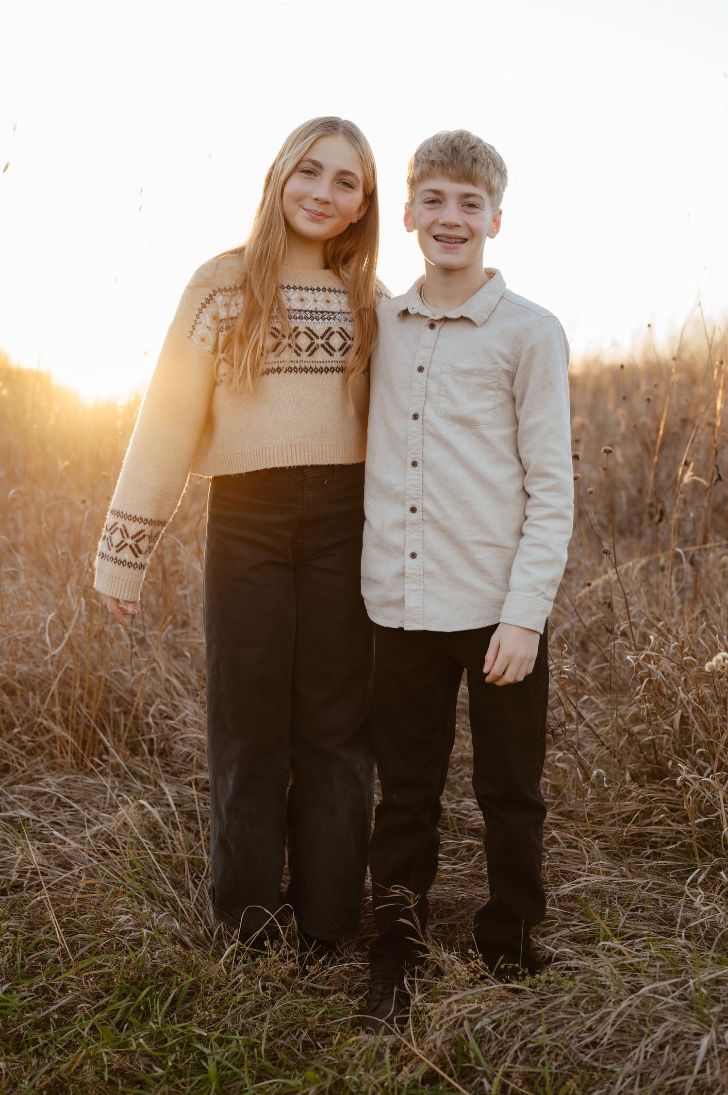 Two smiling teenagers pose together in a golden field at sunset, wearing casual fall outfits.
