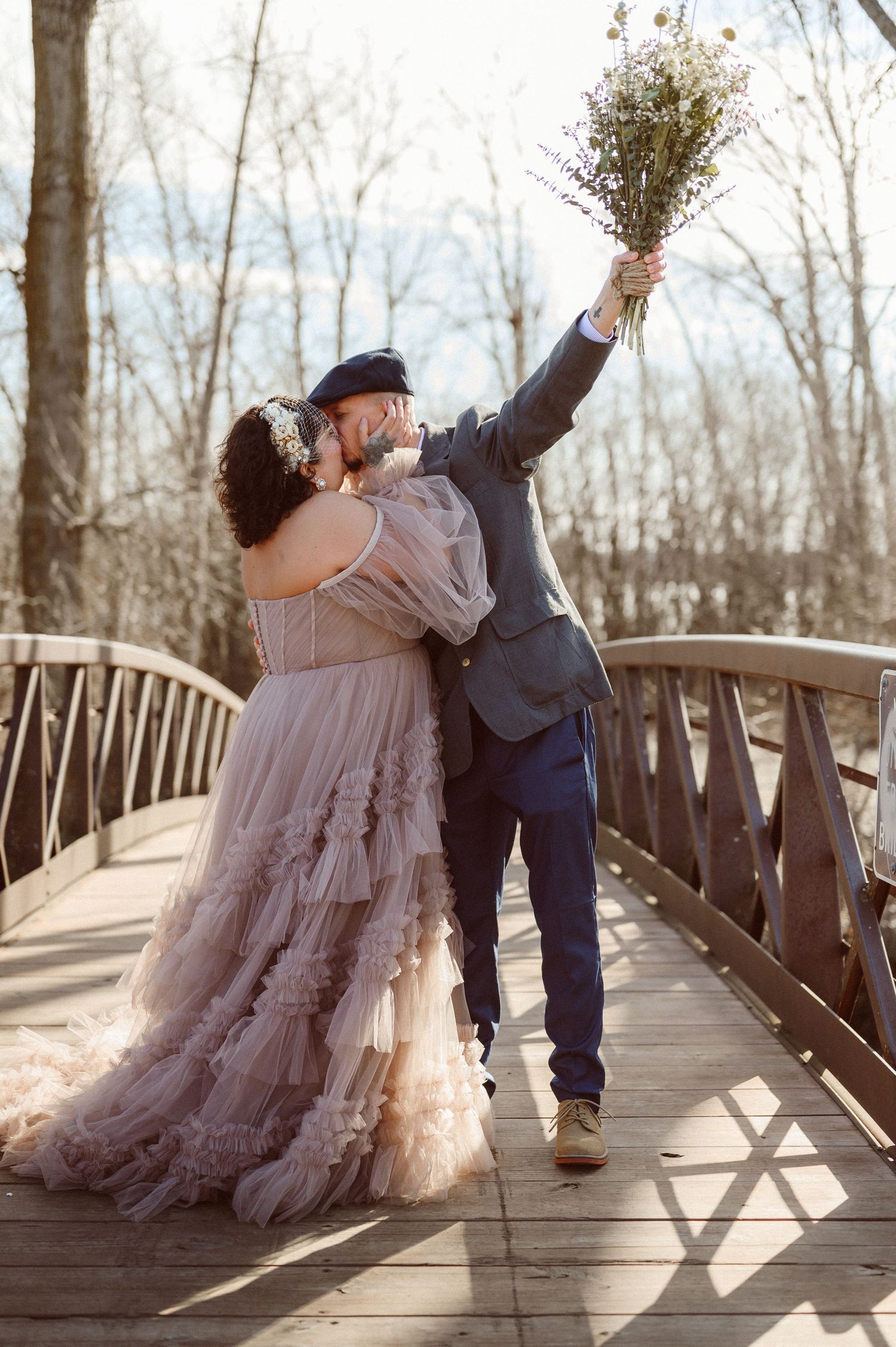 Groom lifting bouquet while kissing bride in a blush tiered tulle gown on a wooden bridge.