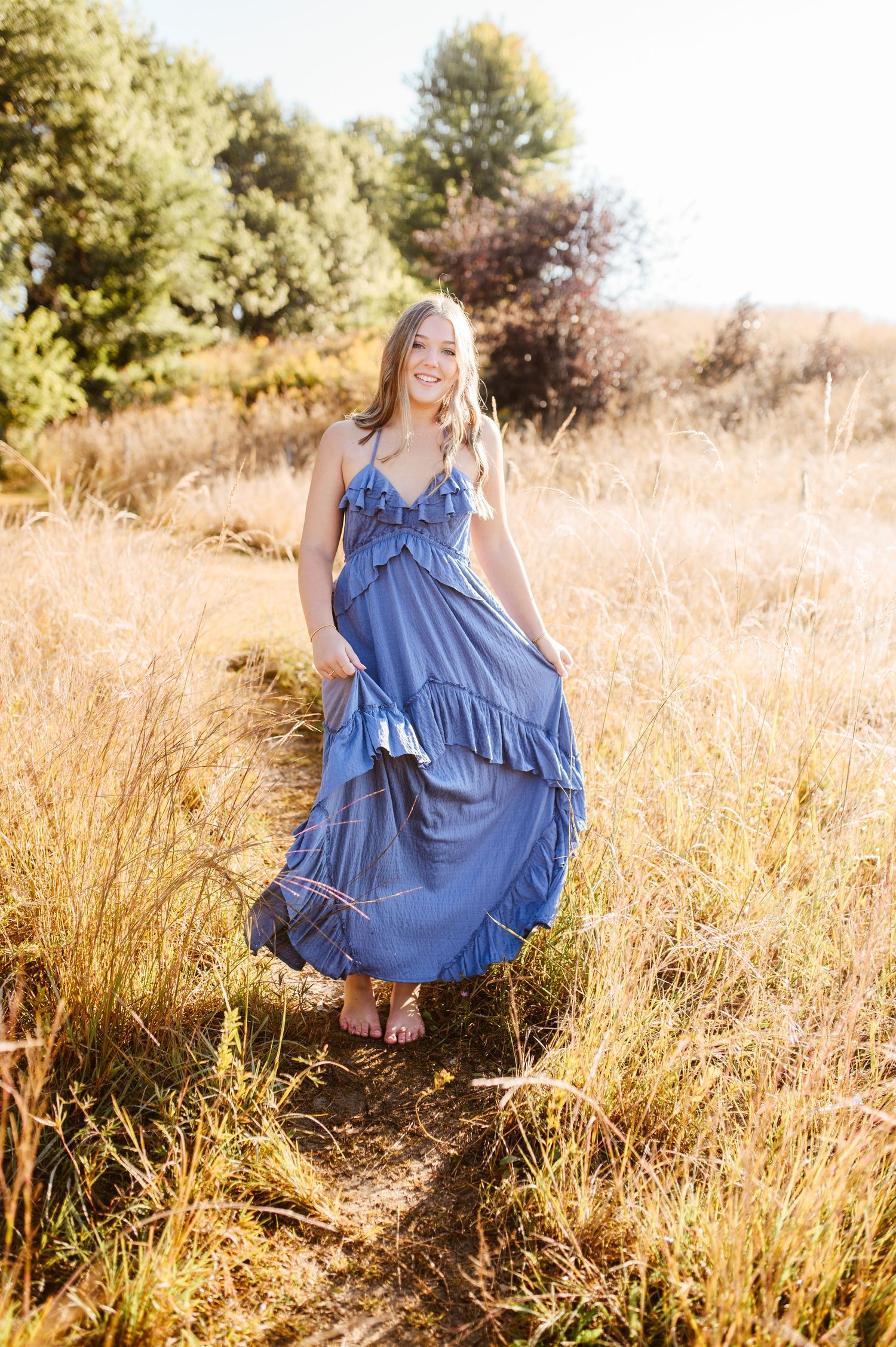 Young woman in a flowing blue maxi dress walking along a path through a golden autumn field.