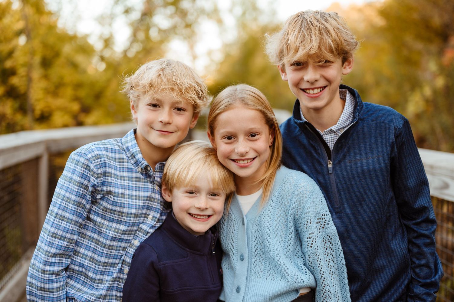 Four blonde siblings smiling together on a wooden bridge surrounded by fall foliage.
