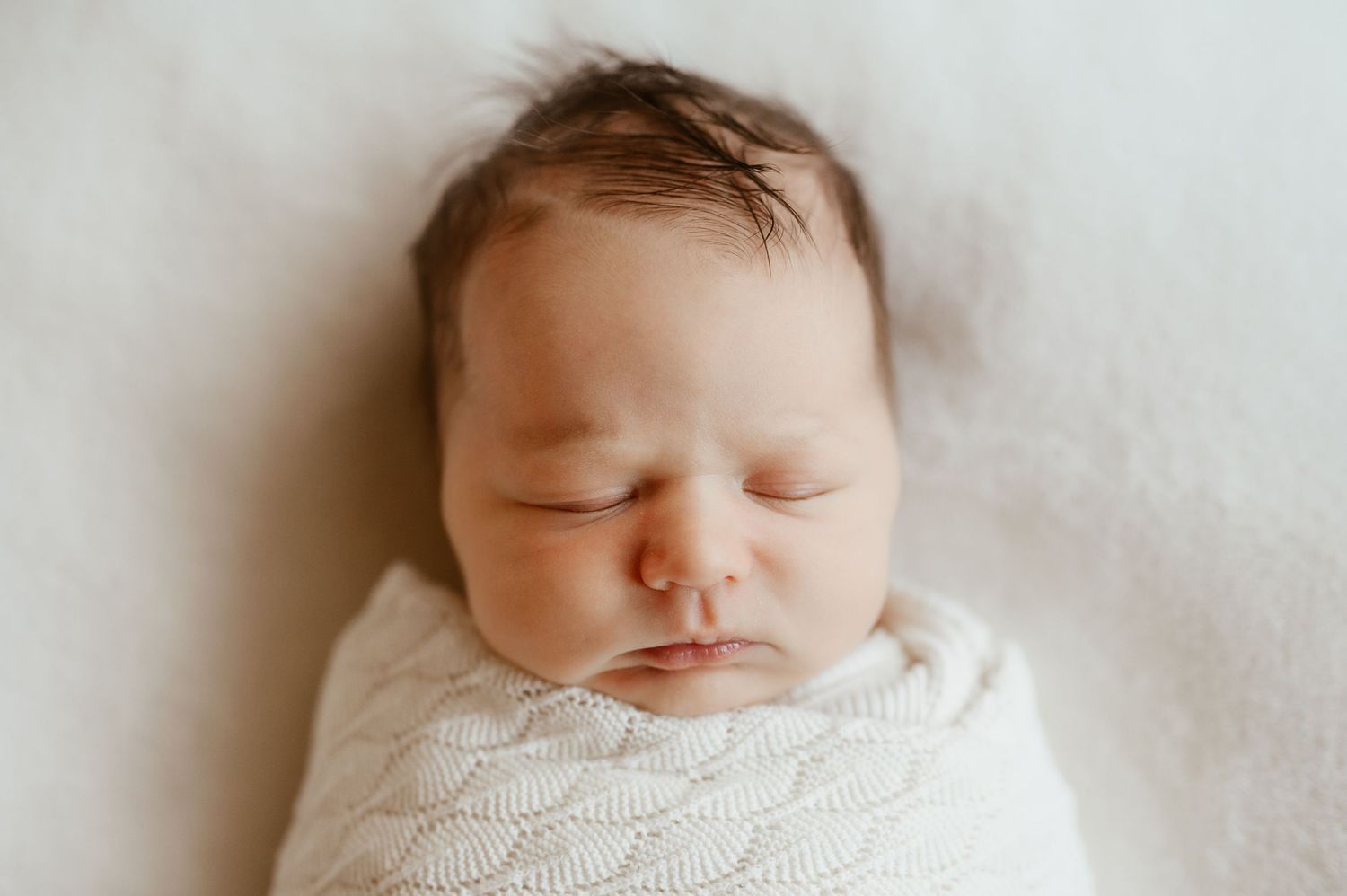A peaceful newborn baby sleeping swaddled in a white knit blanket with dark hair visible.