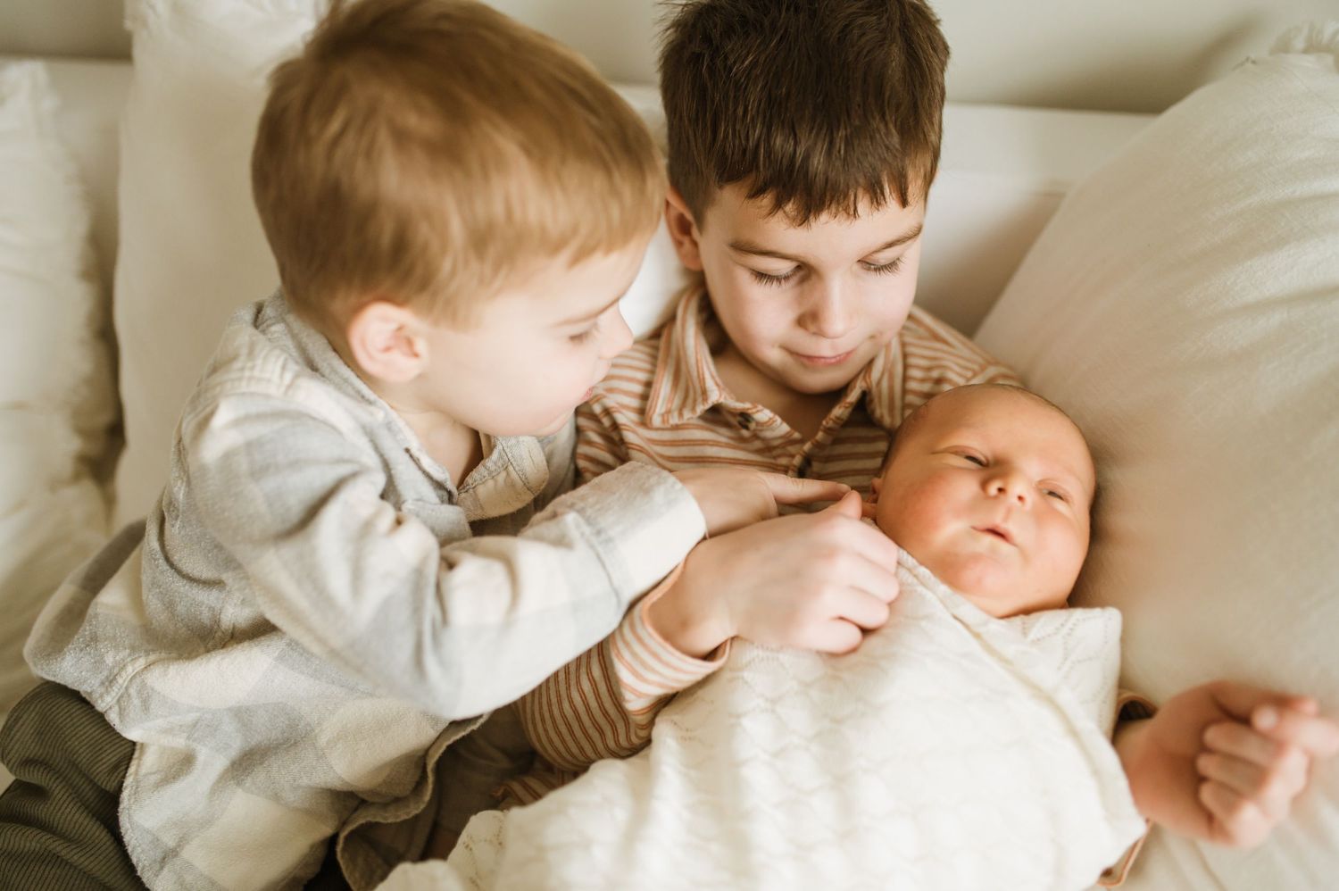 Two young brothers gently touching swaddled newborn sibling on white bed.