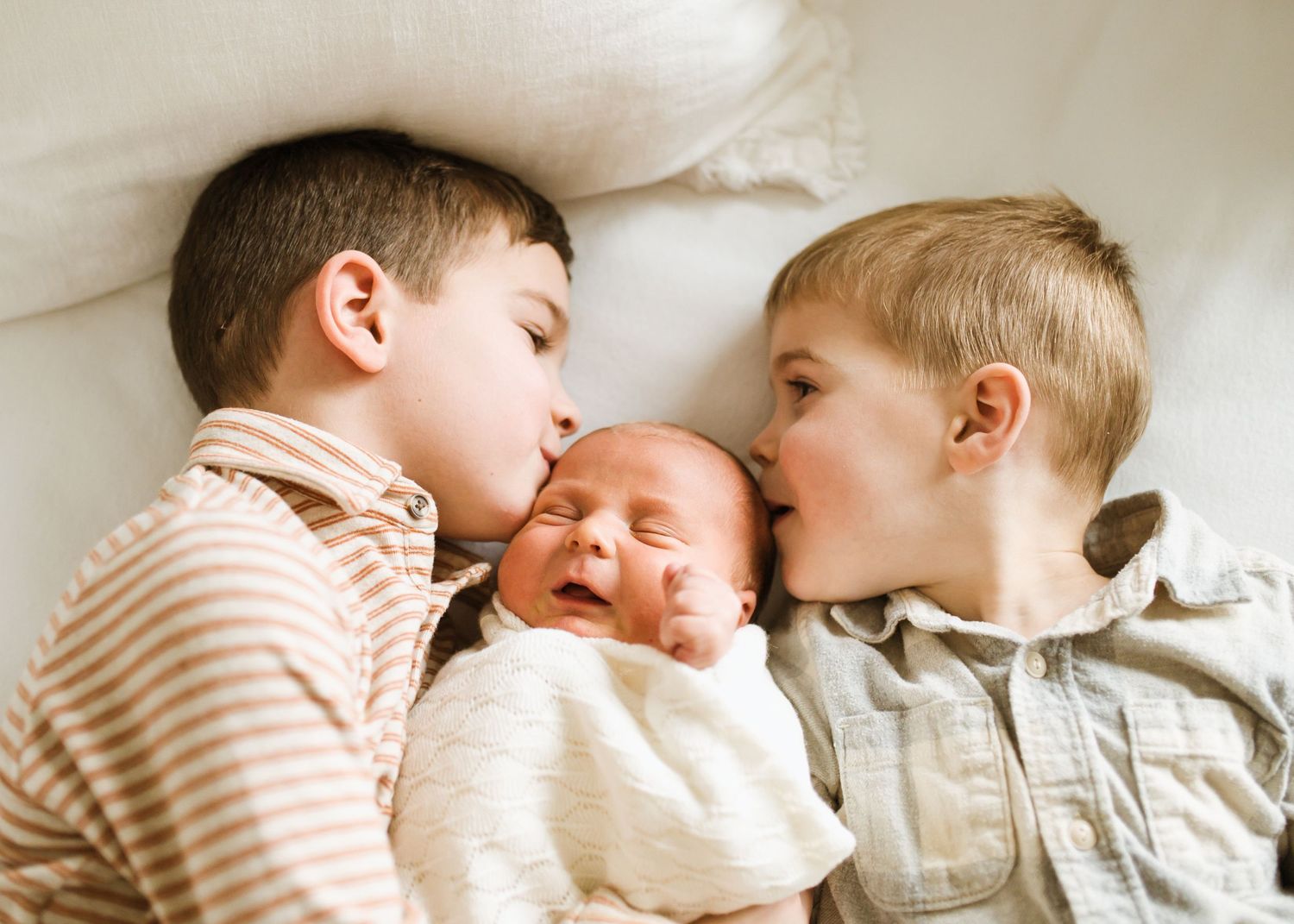 Two young brothers each kiss their newborn sibling on the cheeks while lying in bed.
