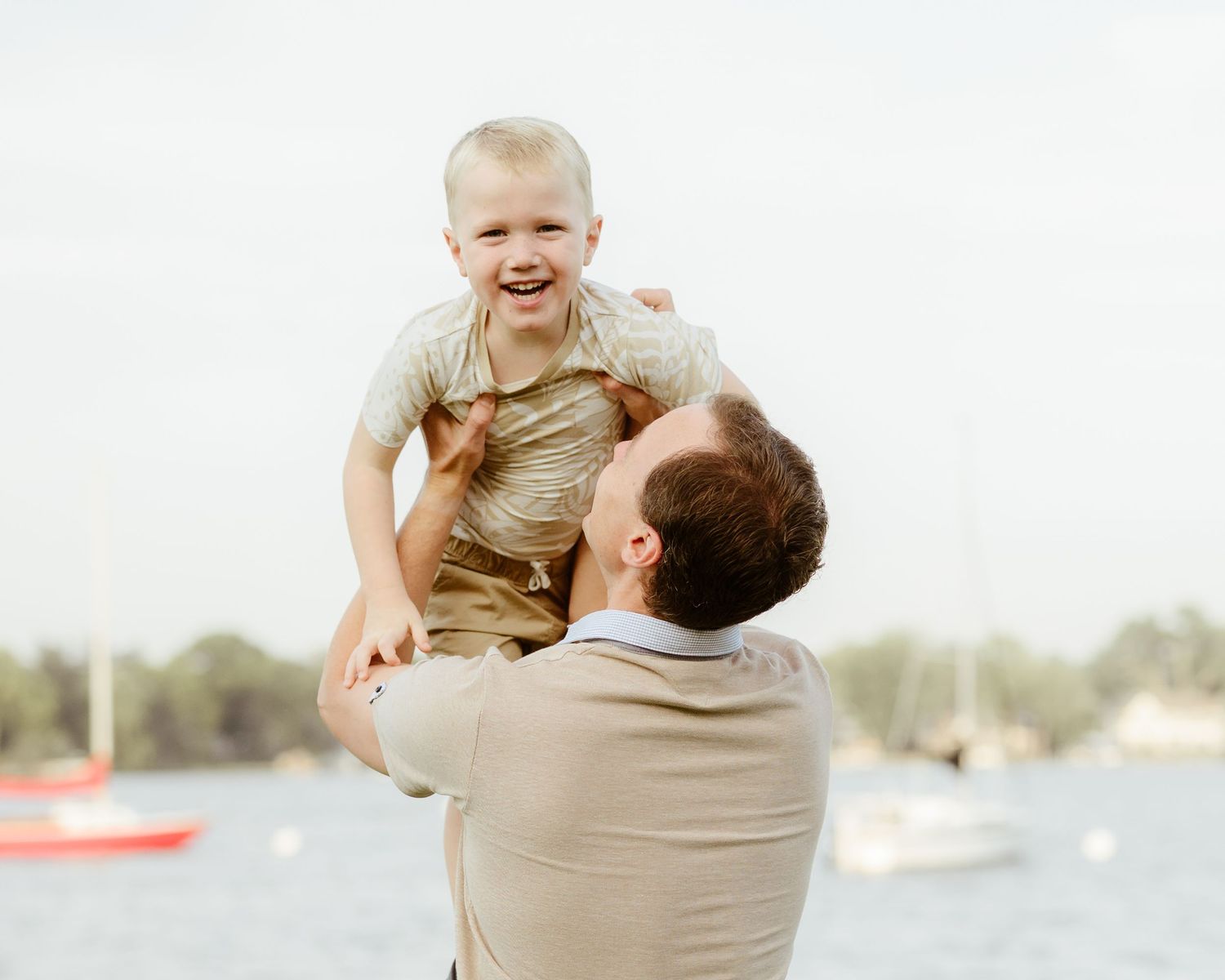 A smiling father lifts his laughing toddler son high in the air near a waterfront.