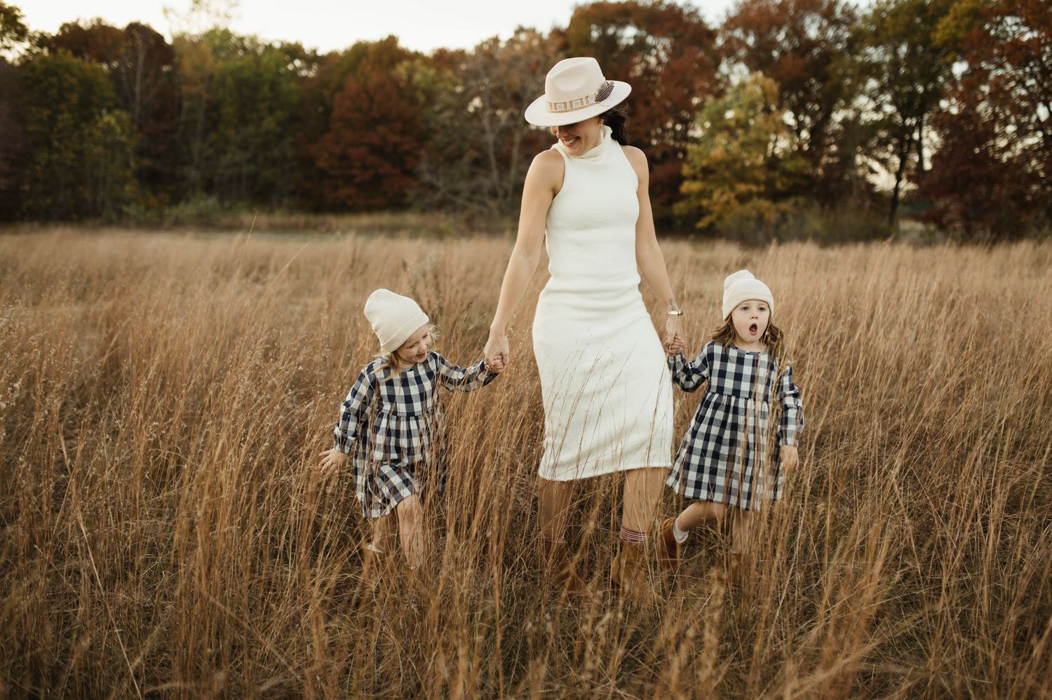 A mother in a white dress and sun hat strolls with two young children in a golden meadow.  Experienced family photographer in Minneapolis