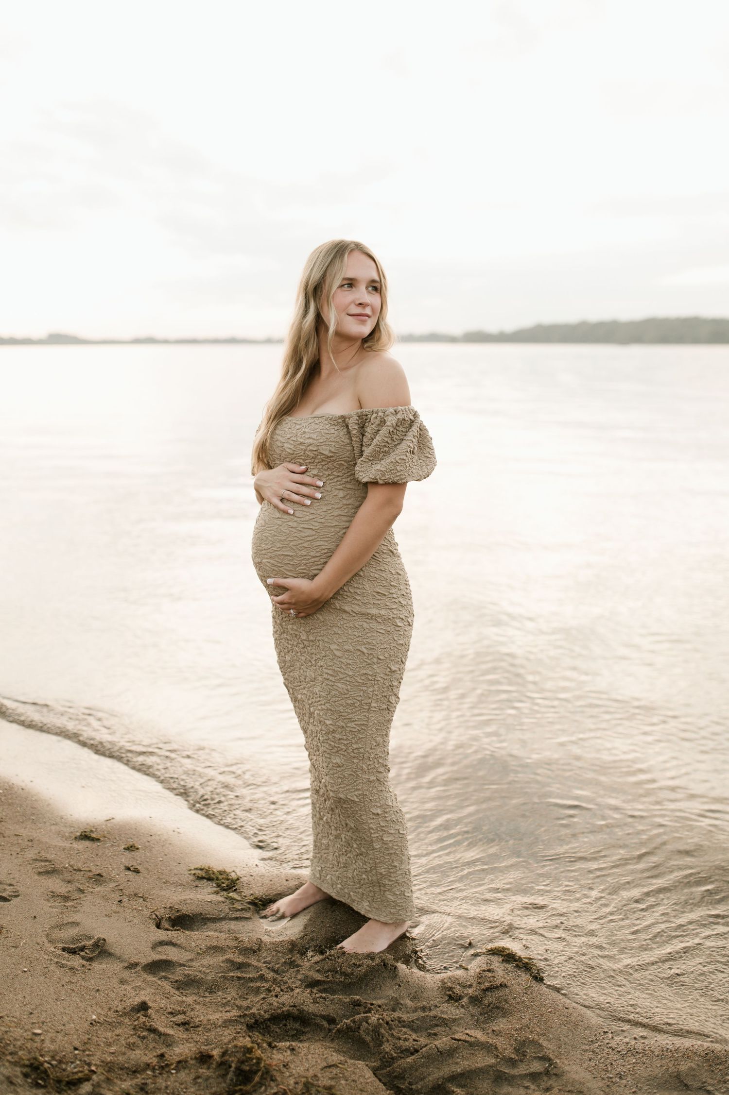 Beautiful pregnant woman in an off-shoulder lace maternity gown standing barefoot on a beach.