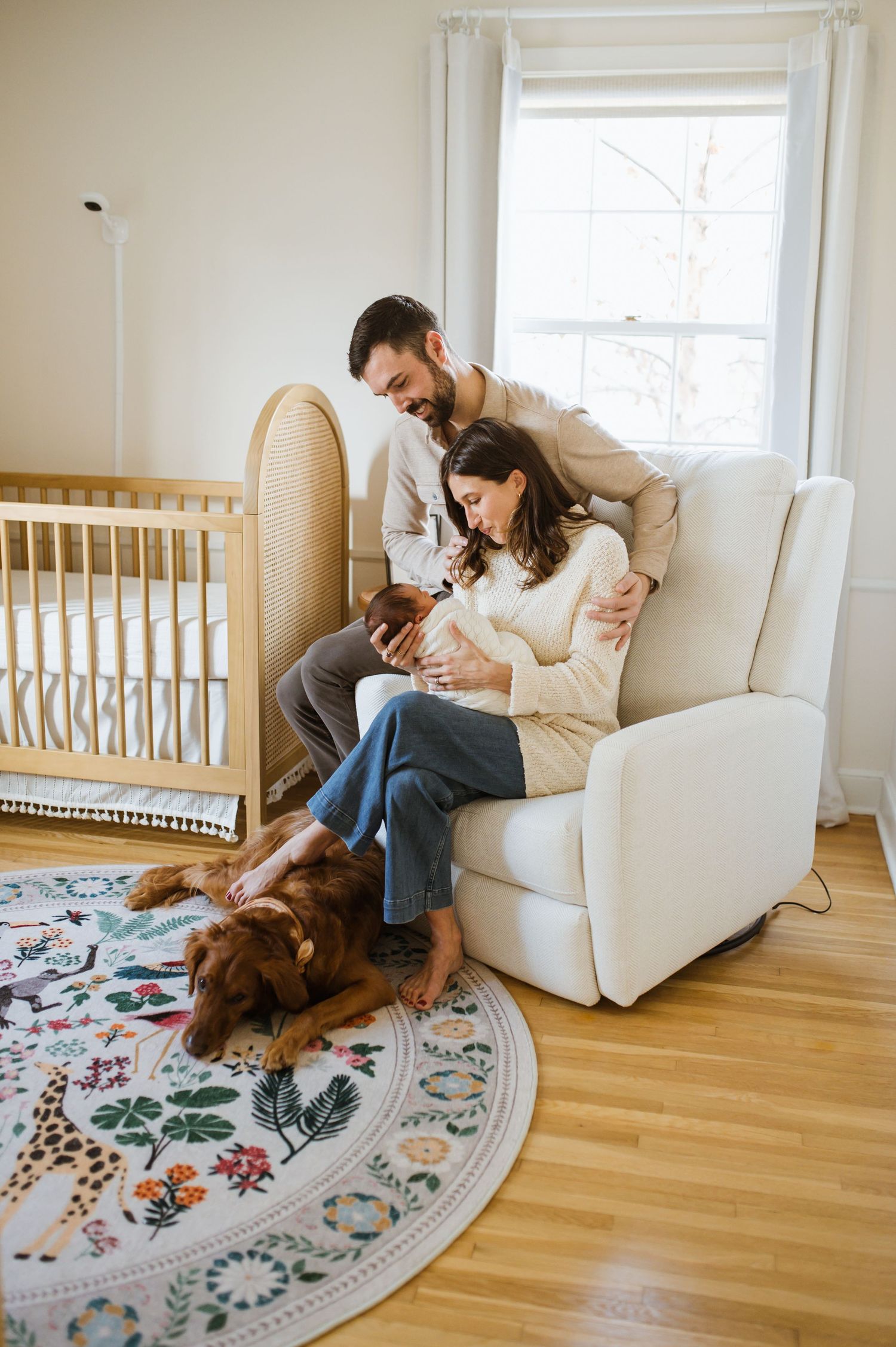 New parents with newborn baby in nursery chair beside crib, with golden retriever resting on floral rug.