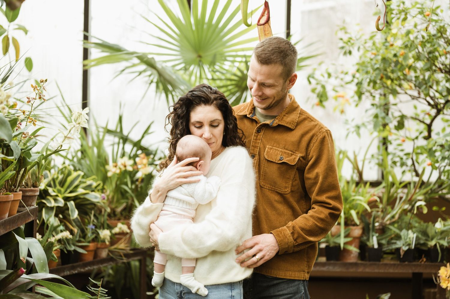 Happy couple with newborn baby among lush green plants in greenhouse.