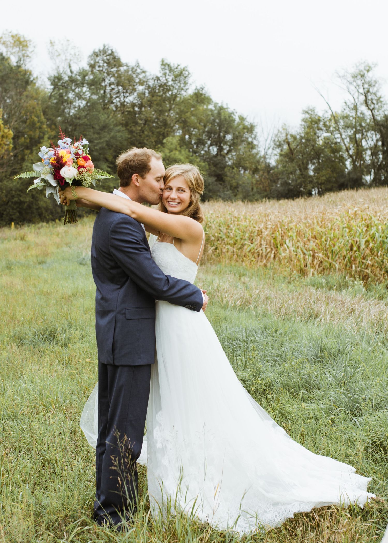 Groom embracing smiling bride holding colorful bouquet in autumn field with cornfield backdrop.