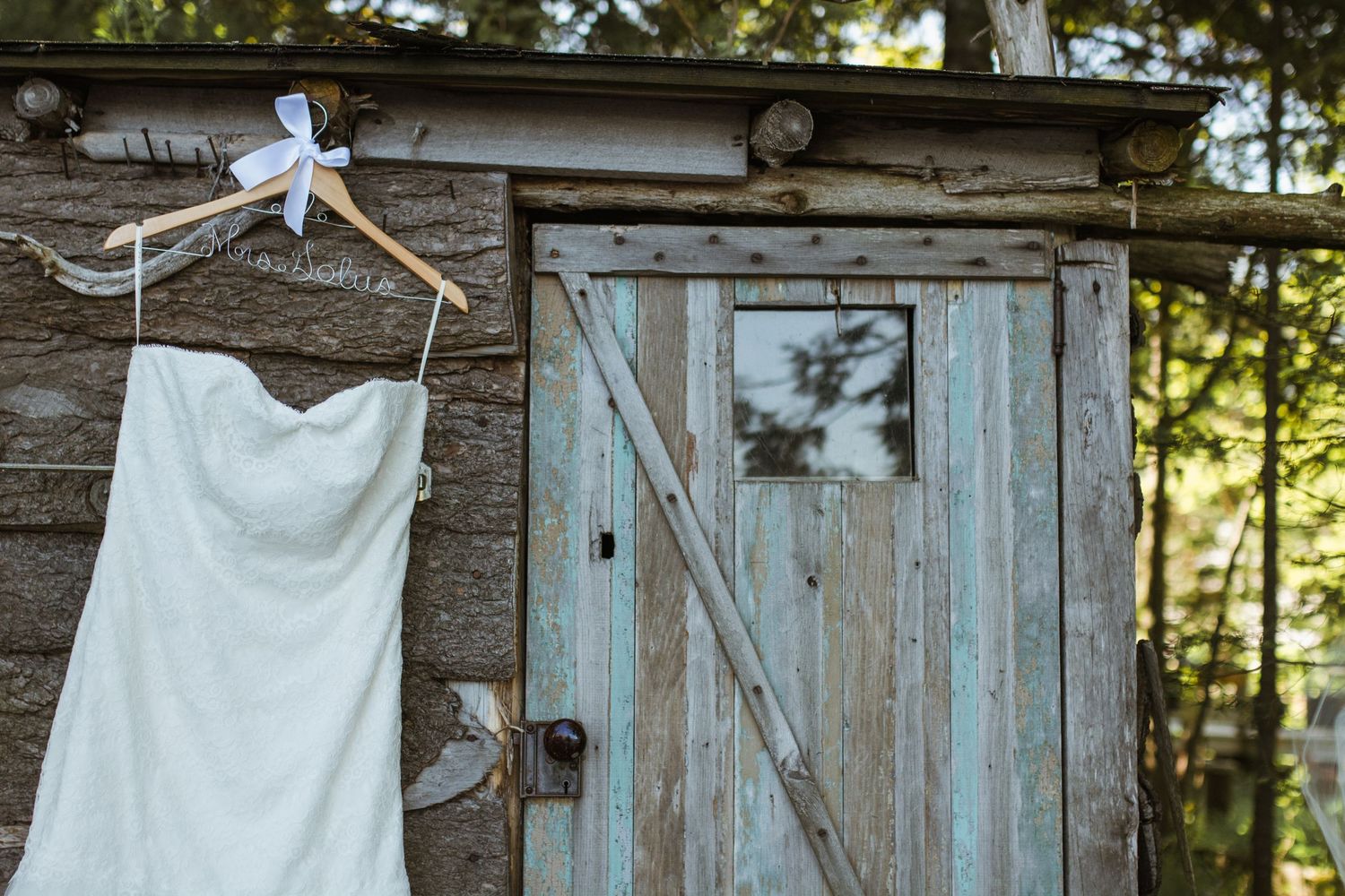 White wedding dress hanging on wooden hanger outside a rustic weathered barn door.