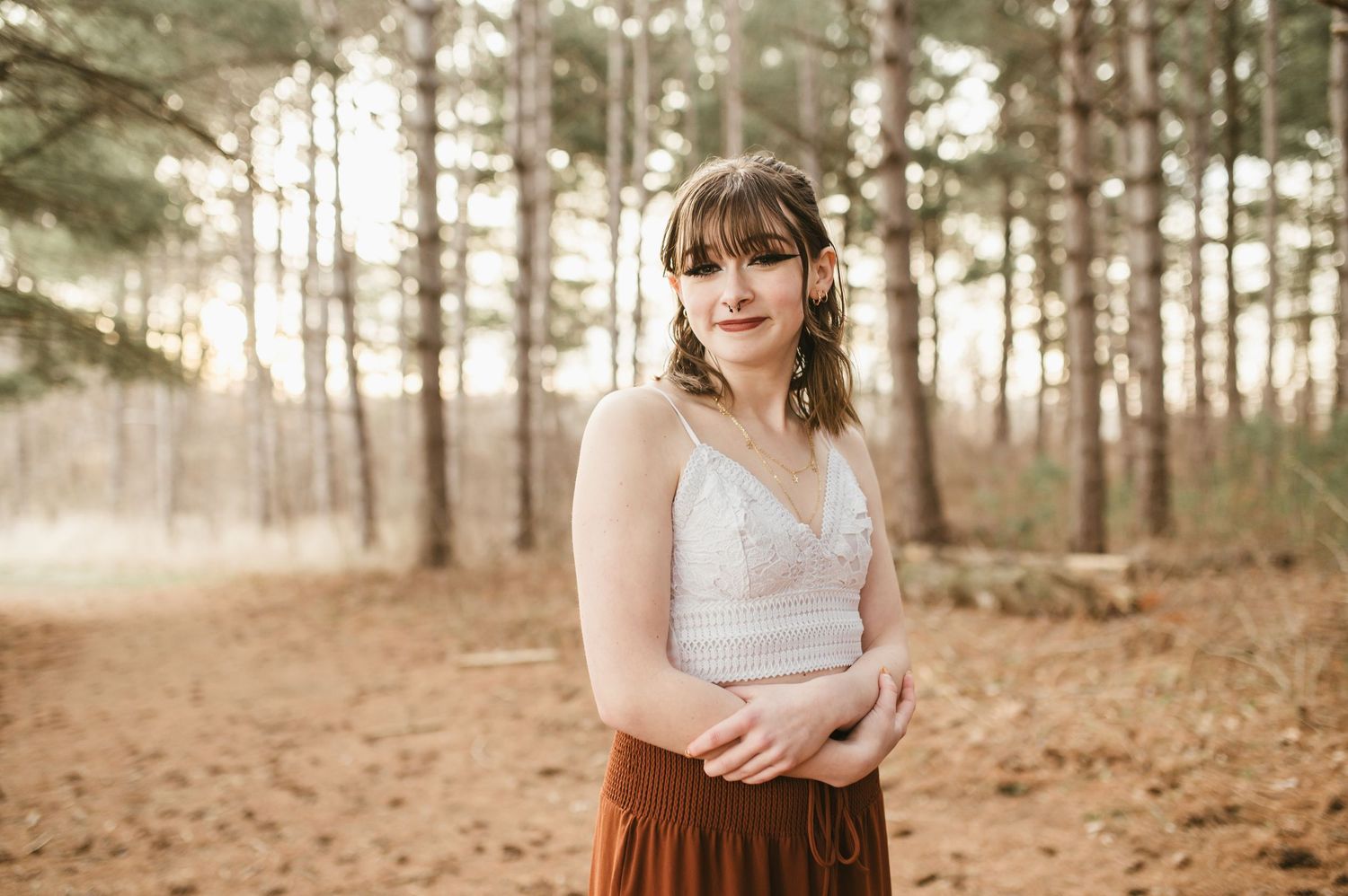 Young woman with floral hair accessory in white lace top and rust skirt poses in misty pine forest.