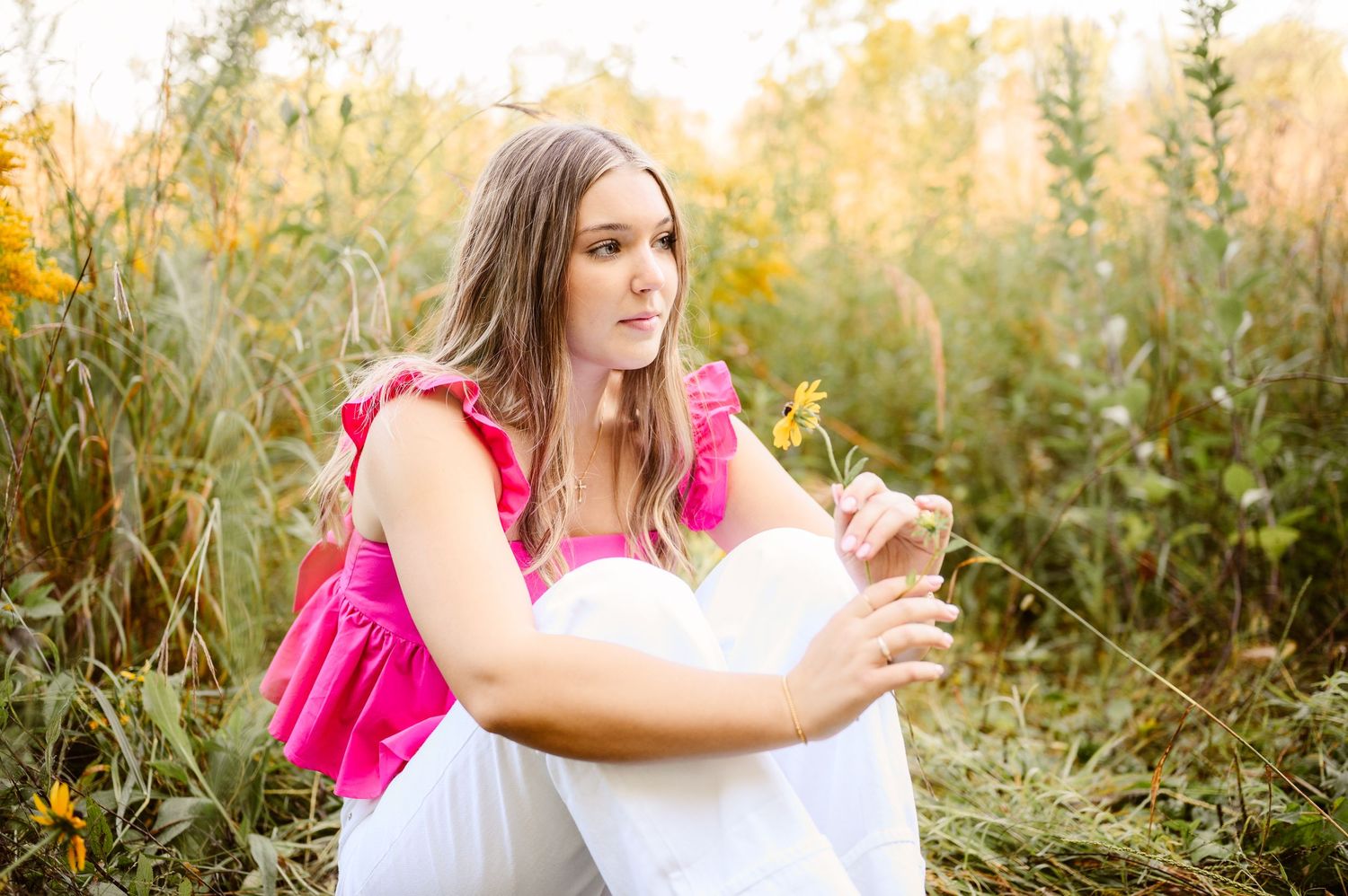Young woman in pink top sits peacefully among wildflowers holding a yellow bloom in golden light. Senior photos twin cities