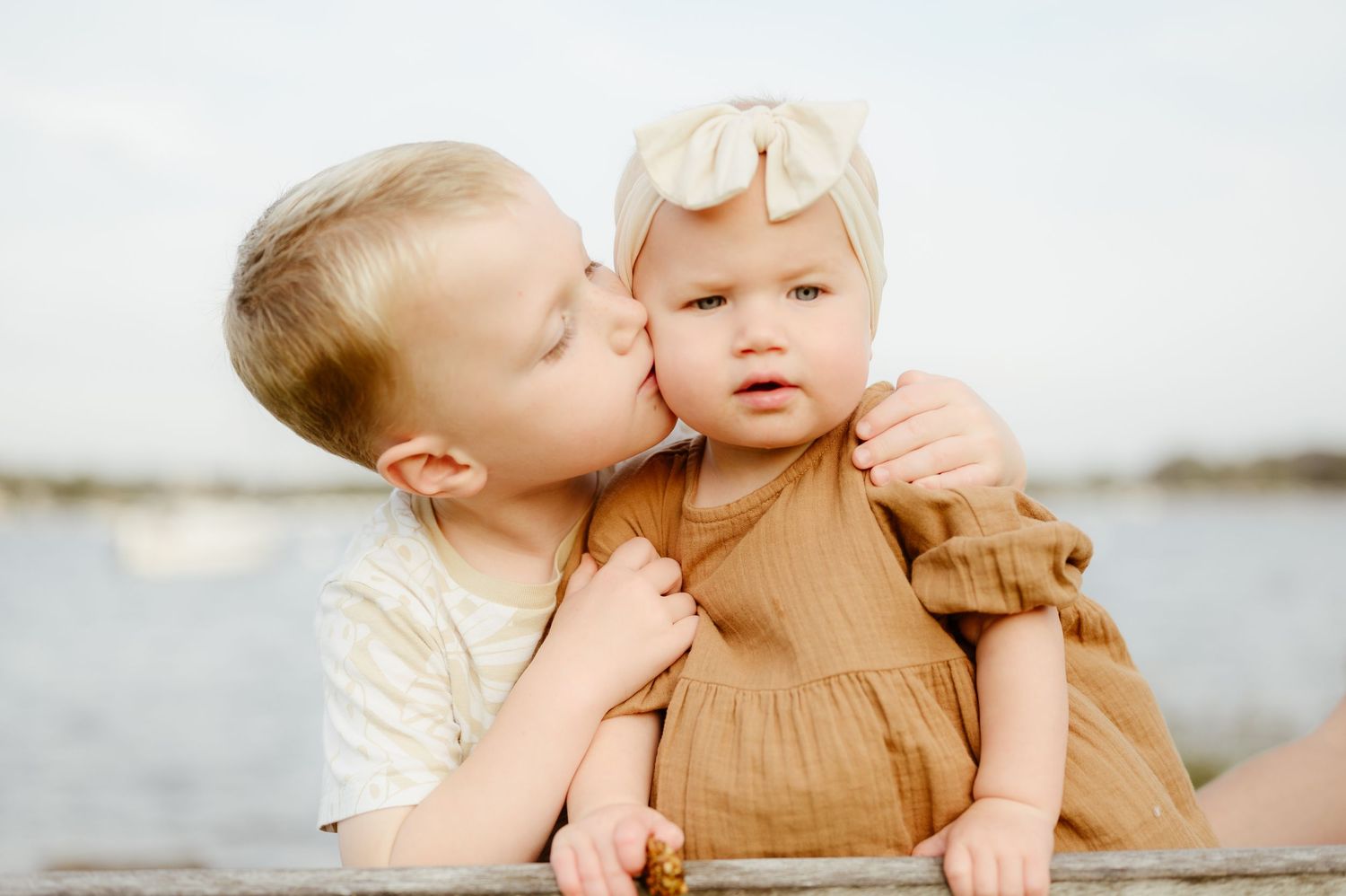 An older brother gently kisses his baby sister on the cheek near a waterfront.