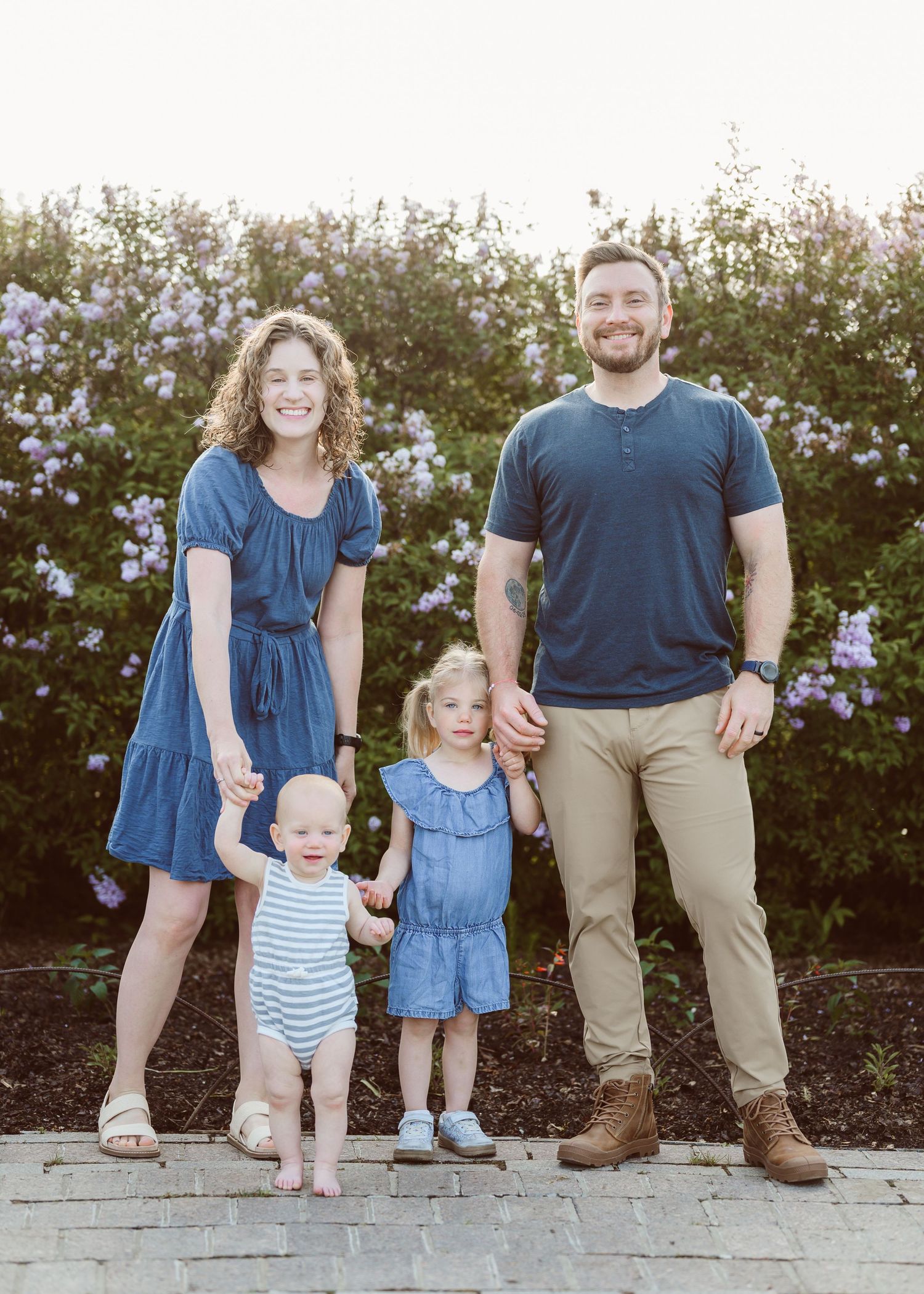 A smiling family of four poses outdoors near blooming bushes, with two young children in matching blue outfits.