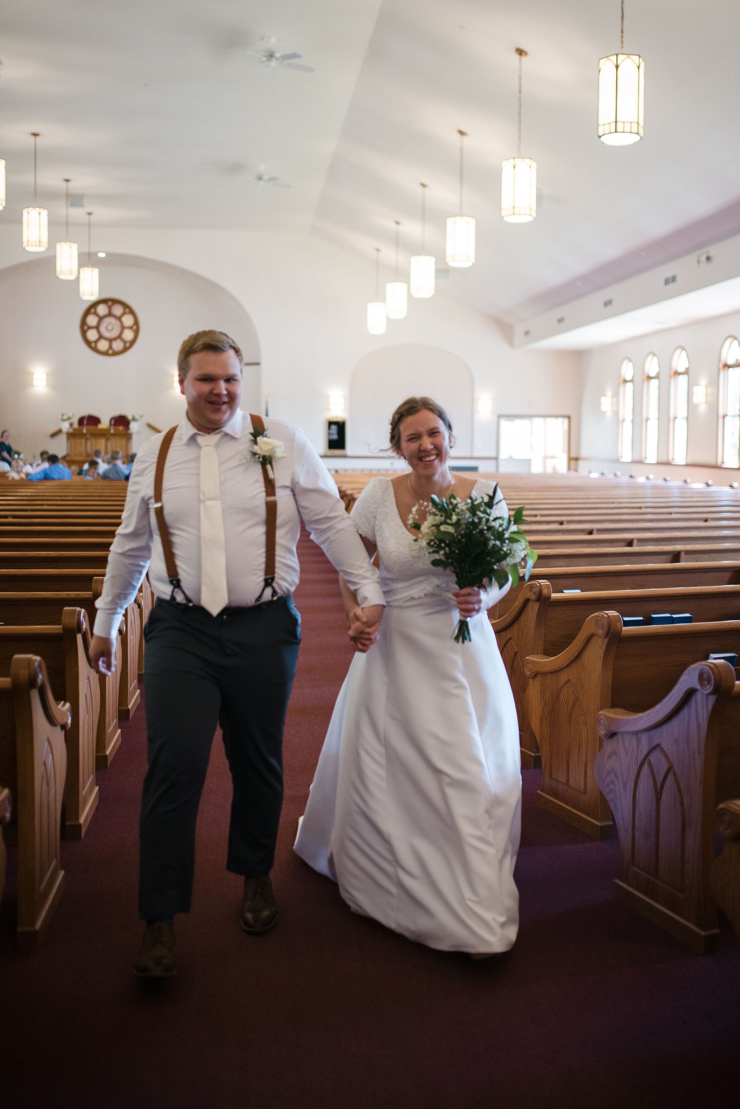 Joyful newlyweds walk down church aisle after ceremony, bride holding greenery bouquet, groom in suspenders.