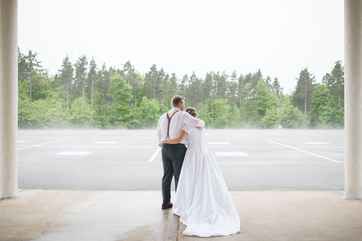 Bride and groom embrace while gazing at misty forest from covered outdoor venue on wedding day.