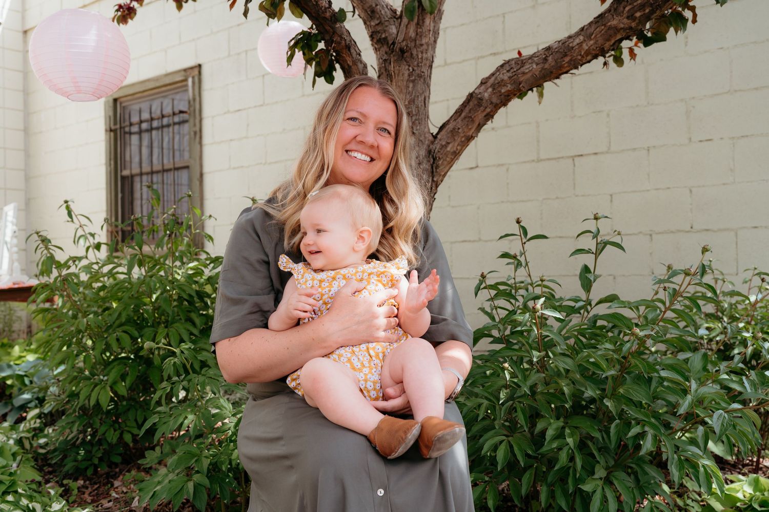 A smiling mother holds her laughing baby girl outdoors near a pink balloon. Meet your Minneapolis Newborn Photographer