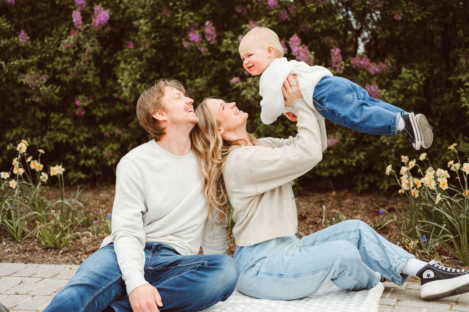 A happy young family sits outdoors, with a smiling mother lifting a laughing baby while father sits beside them.
