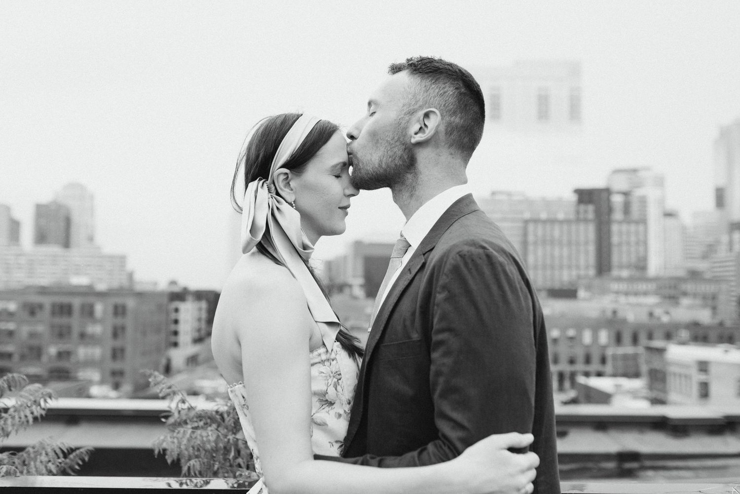 Black and white photo of a couple sharing a kiss on a rooftop with city skyline behind them.