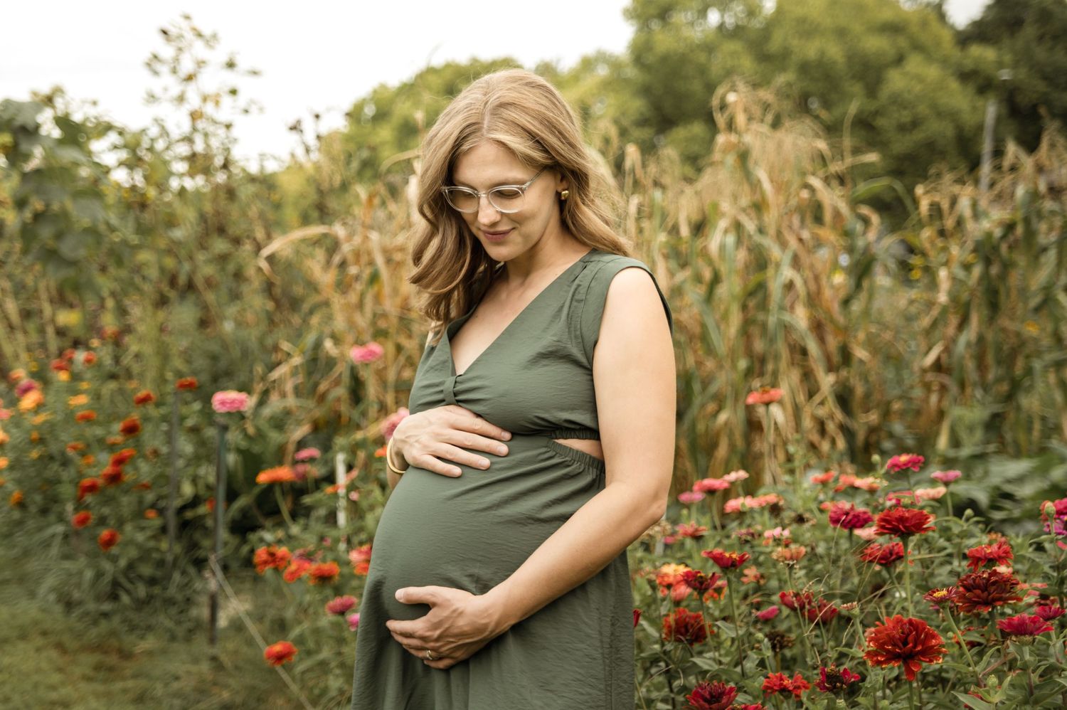 Pregnant woman in olive green dress lovingly cradling baby bump surrounded by colorful flowers in garden.