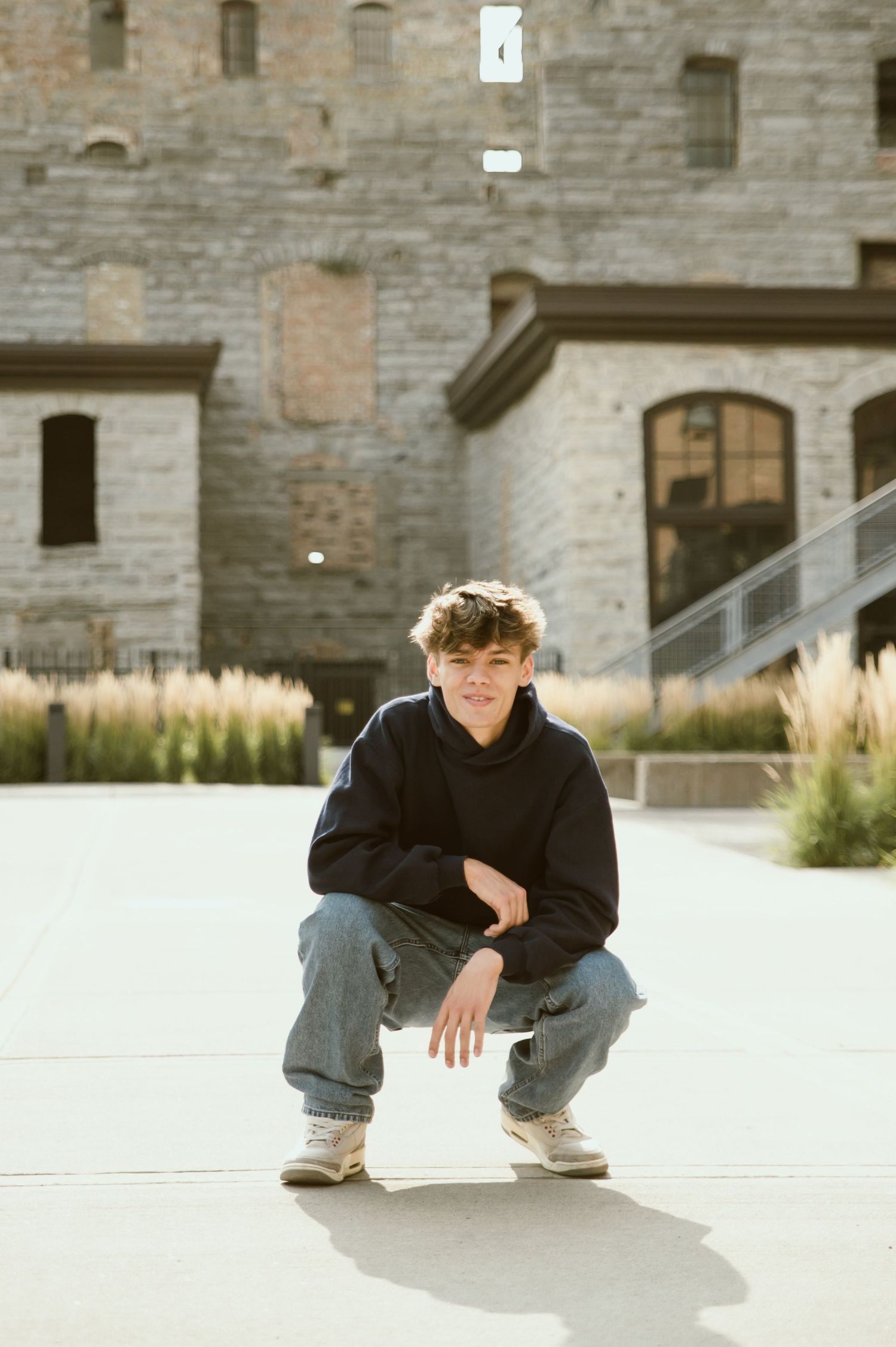 Teen boy in black hoodie and jeans crouching in front of a stone building outdoors.