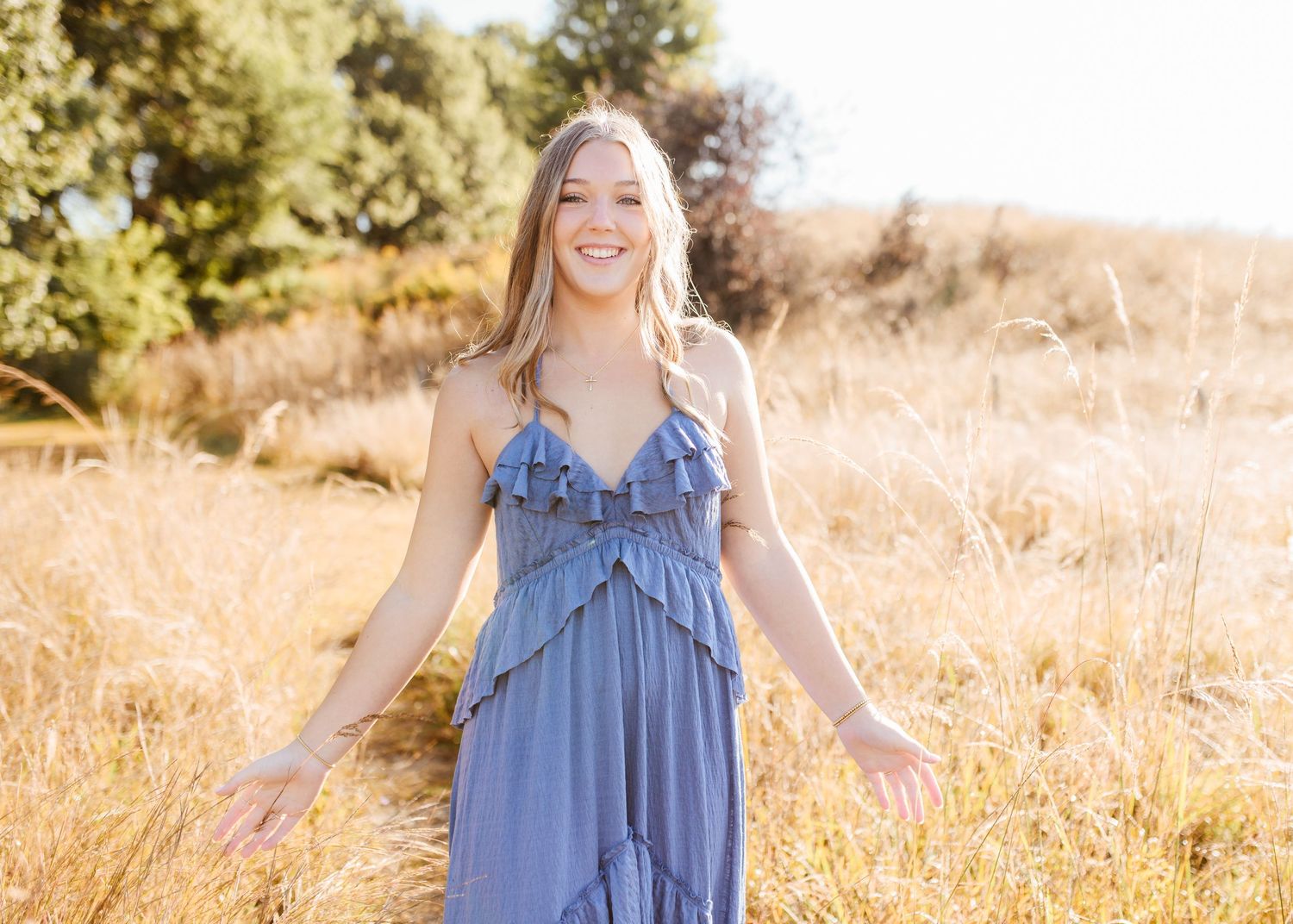 Smiling young woman in a blue ruffled maxi dress standing in a sunlit golden field with arms outstretched.