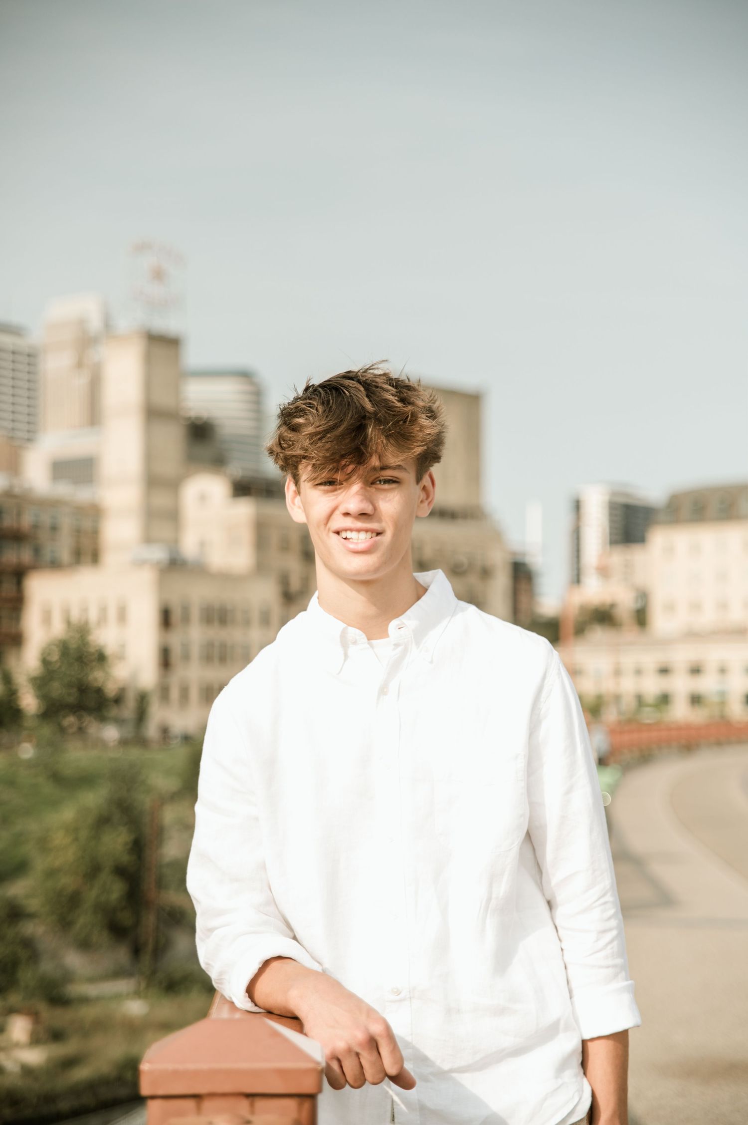 Smiling teen boy in white linen shirt posing on rooftop with sunny city skyline behind him.