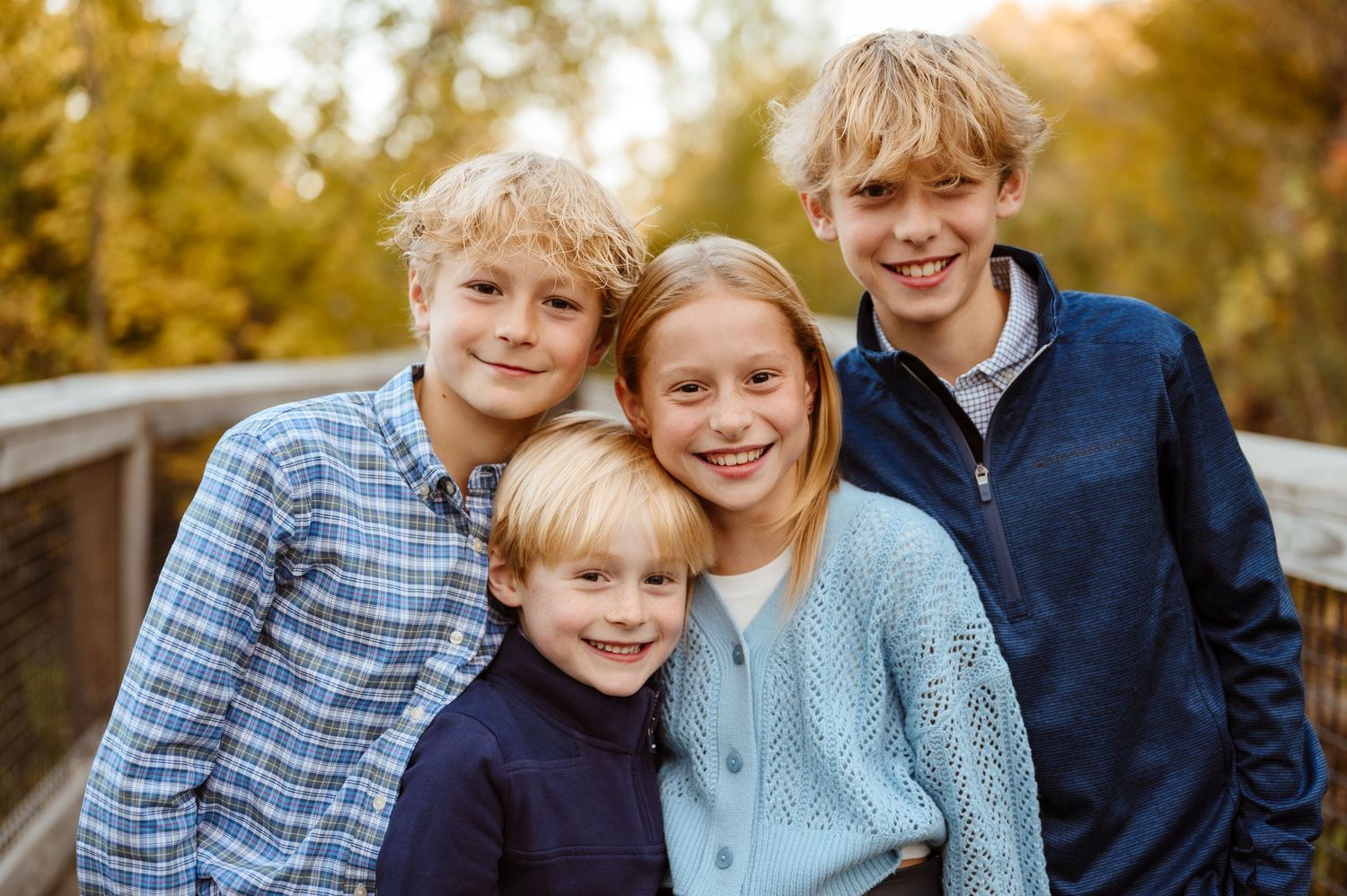 Four blonde siblings smiling together on a wooden bridge in autumn.