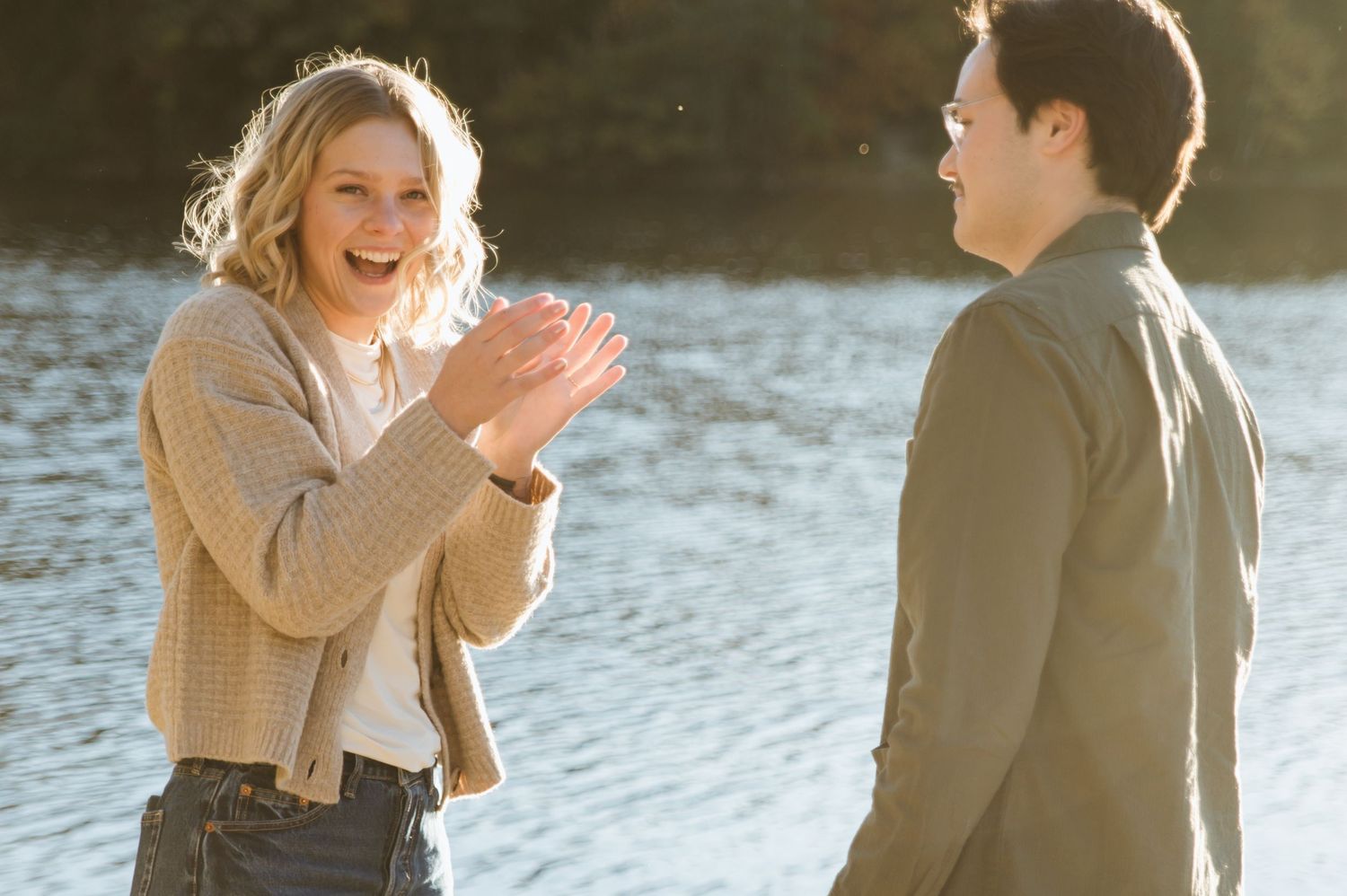 A joyful woman clapping her hands laughs with a man by a sunlit lake during an outdoor engagement session.