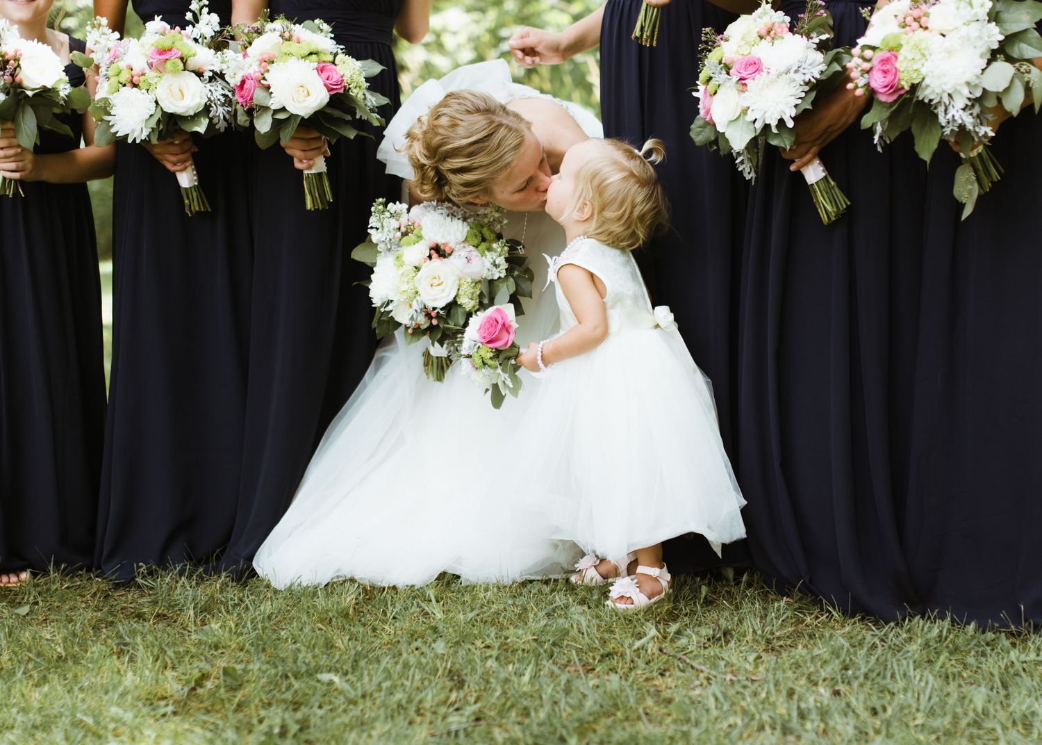 Bride kissing flower girl in white dress surrounded by bridesmaids in navy gowns holding pink and white bouquets.