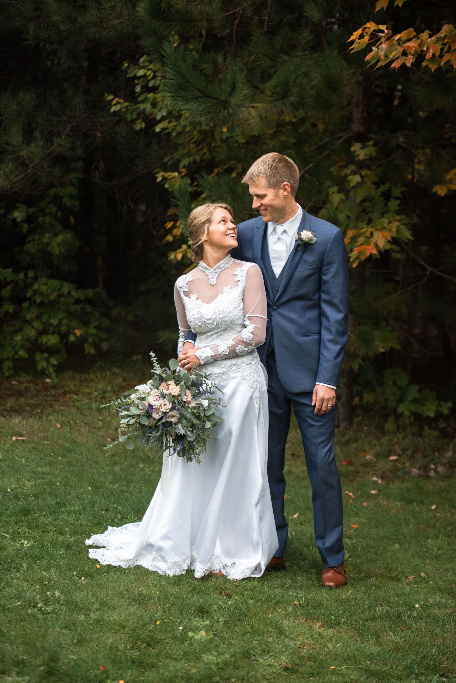 Bride and groom embrace on green lawn with lush forest background, bride holding greenery bouquet.