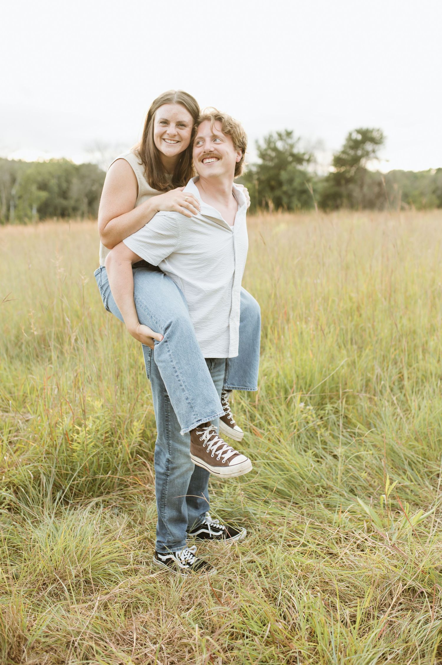 A smiling man gives a laughing woman a piggyback ride in a tall grass field outdoors.