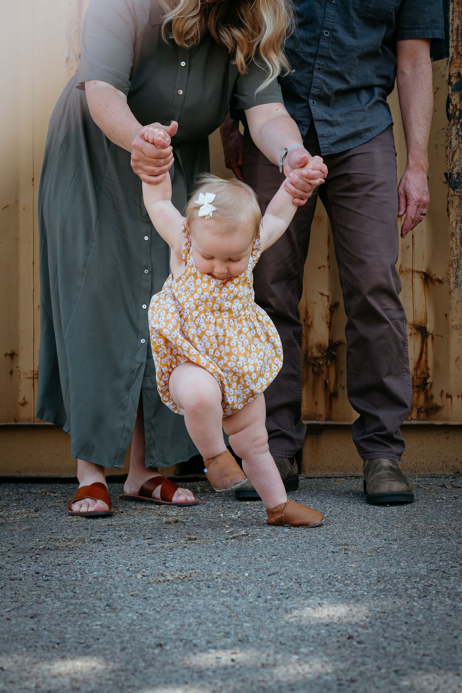 Parents help their baby girl take her first steps outdoors in a floral romper. St. Paul Family Photographer.