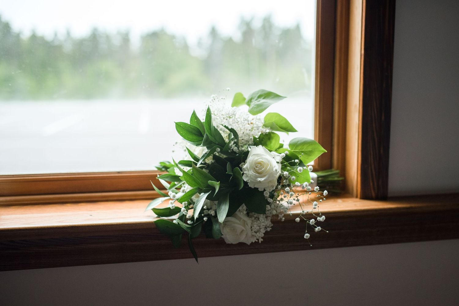 Small white rose and greenery bridal bouquet resting on wooden windowsill with soft natural light.