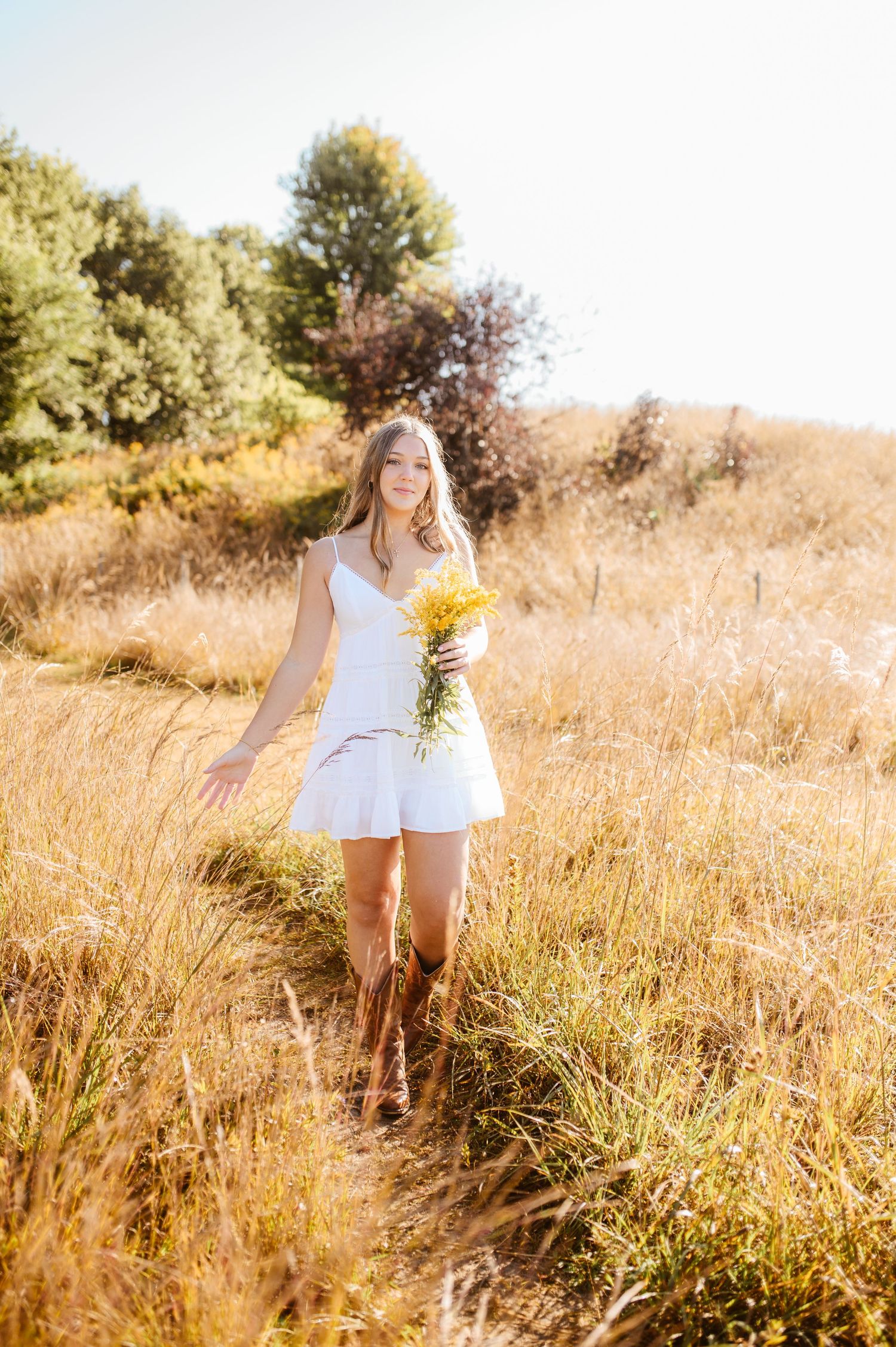 Young woman in white dress and cowboy boots walks through golden field holding yellow wildflowers on sunny day.
