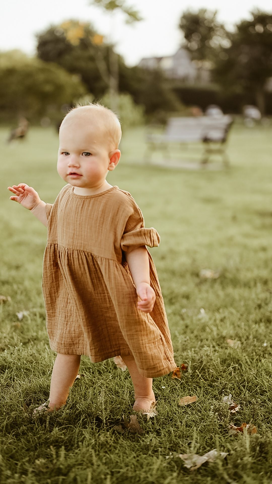 Cute baby girl in a brown dress walks barefoot on green grass in a sunny park. Minneapolis motherhood photographer
