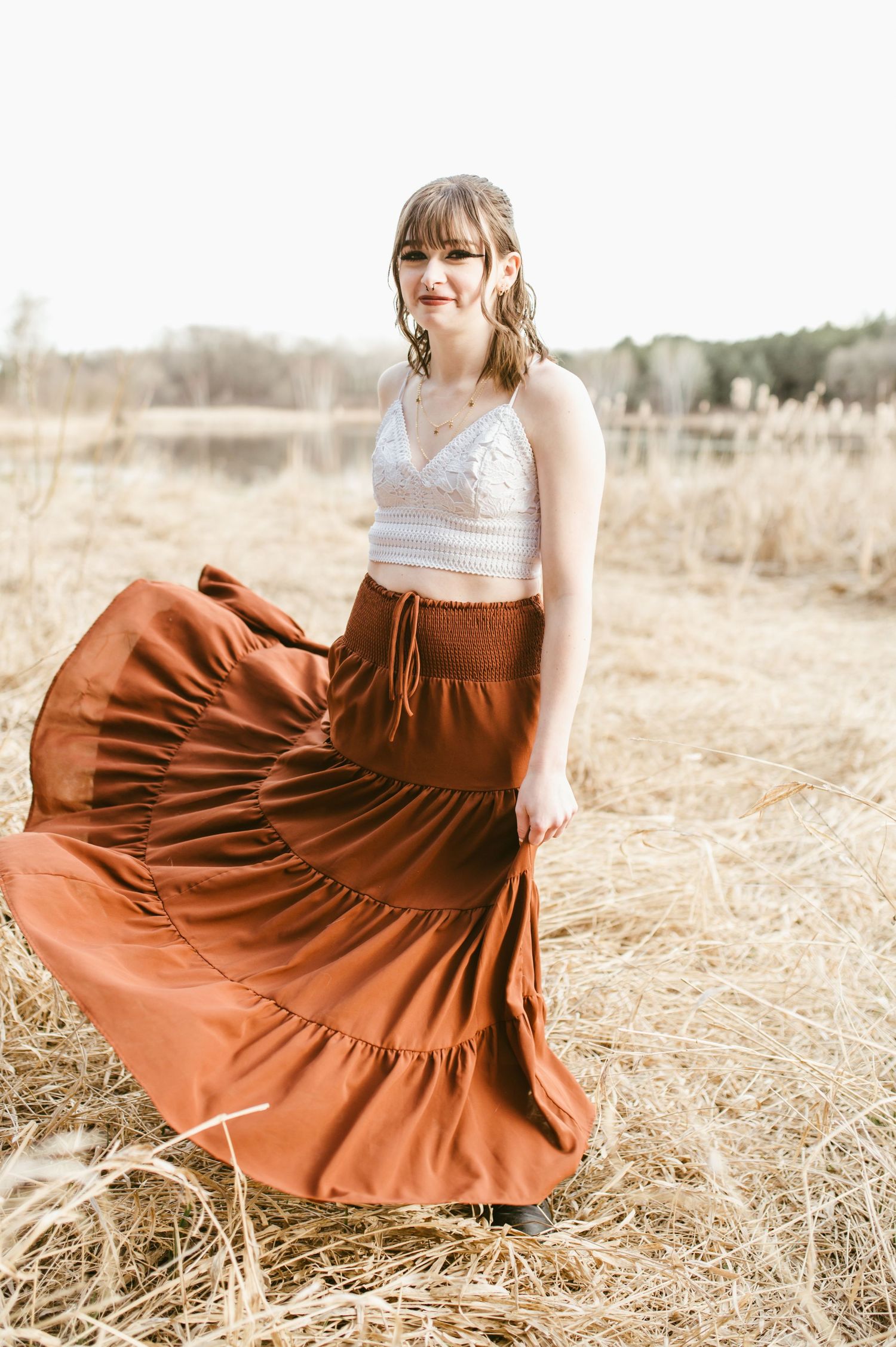 Young woman twirls rust-colored tiered maxi skirt with white lace top in golden dried grass field.