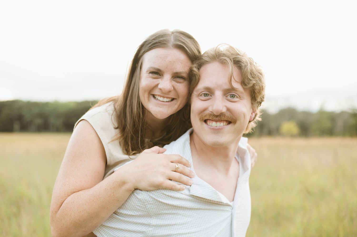 A smiling couple poses together in a golden meadow, the woman hugging the man from behind happily.