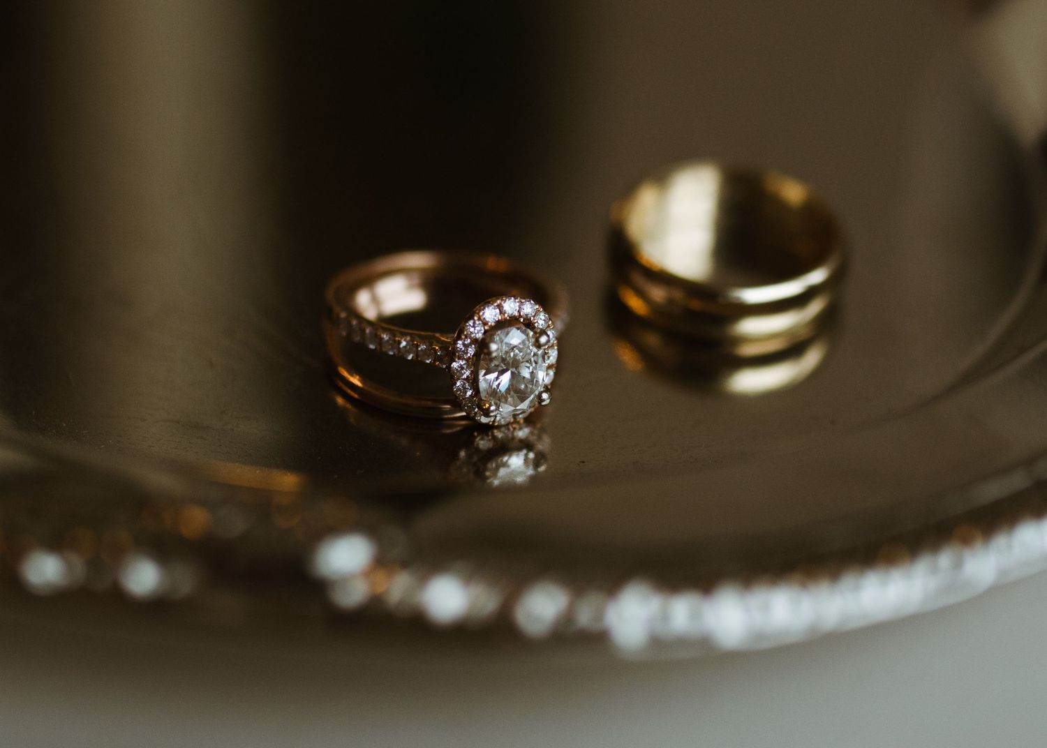 Close-up of diamond halo engagement ring and gold wedding band displayed on decorative silver tray.