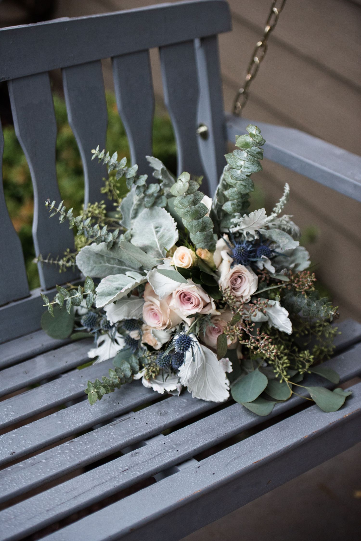 Romantic bridal bouquet with blush roses, eucalyptus, and dusty miller resting on a gray wooden bench.