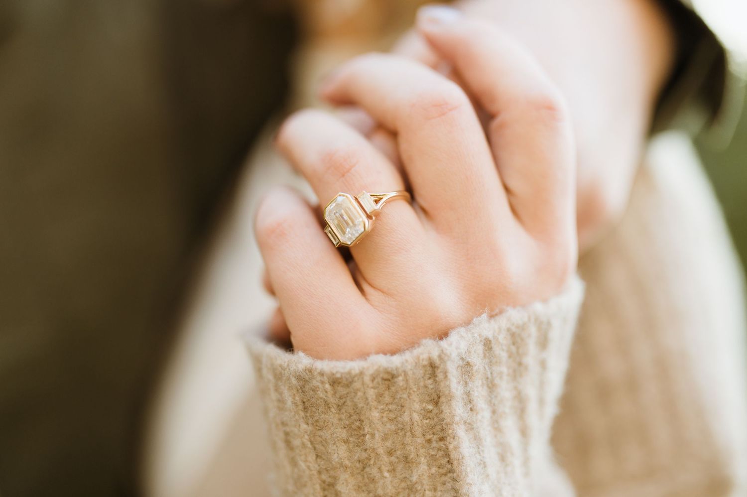 Close-up of a gold emerald-cut engagement ring on a woman's hand wearing a cream knit sweater.