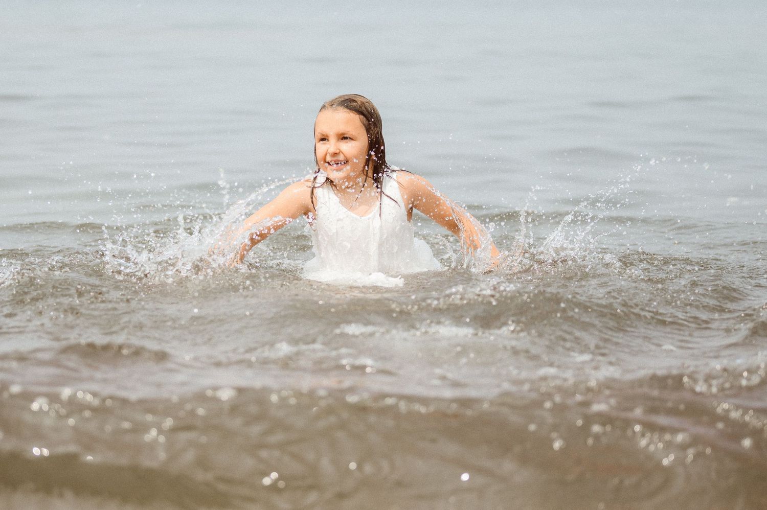 A smiling girl in a white dress splashes and plays in ocean waves at the beach.