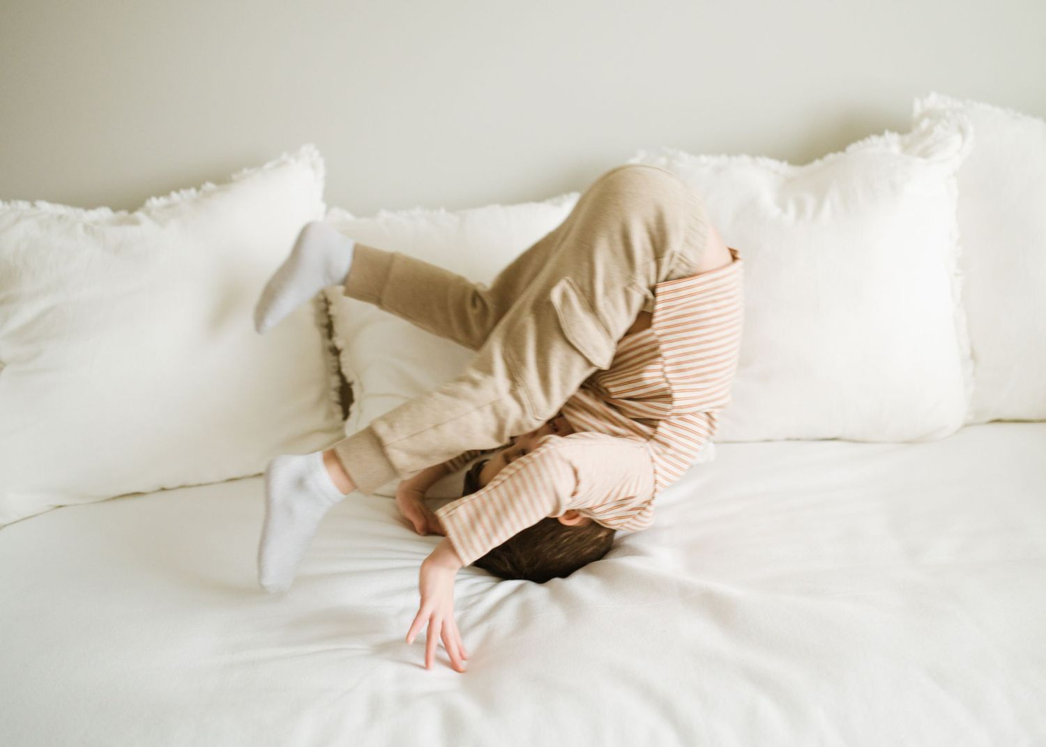 Young child playfully hiding face-down among white pillows on a bed.