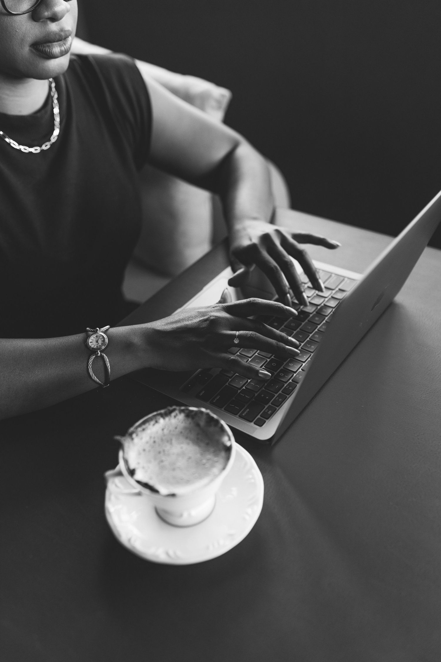 Black and white photo of woman typing on laptop with coffee cup on table.